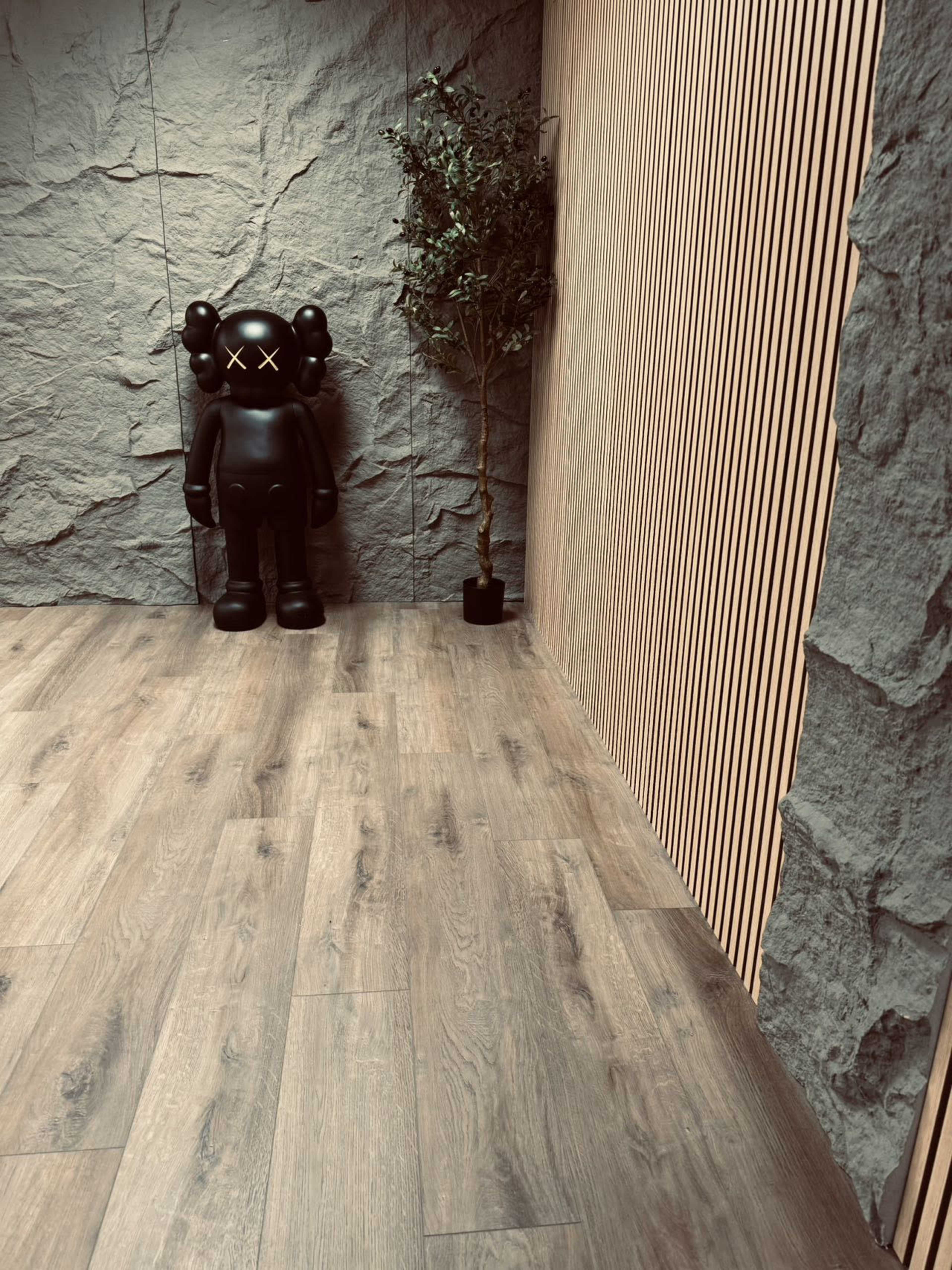 A black figurine stands against a textured stone wall next to a small potted plant in a minimalist indoor space featuring wooden flooring.