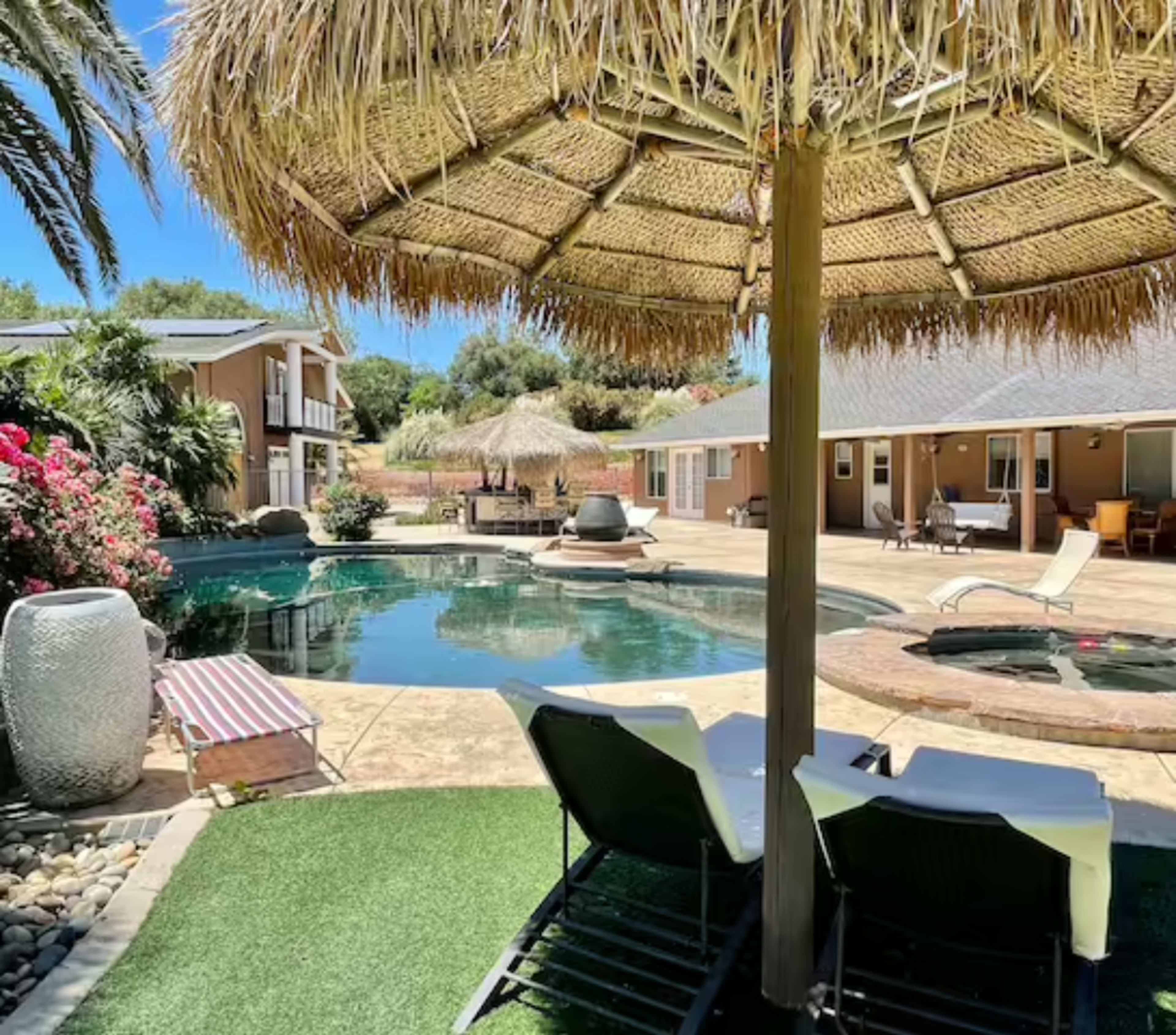 A pool area surrounded by tropical landscaping, featuring lounge chairs, a thatched umbrella, and a hot tub.