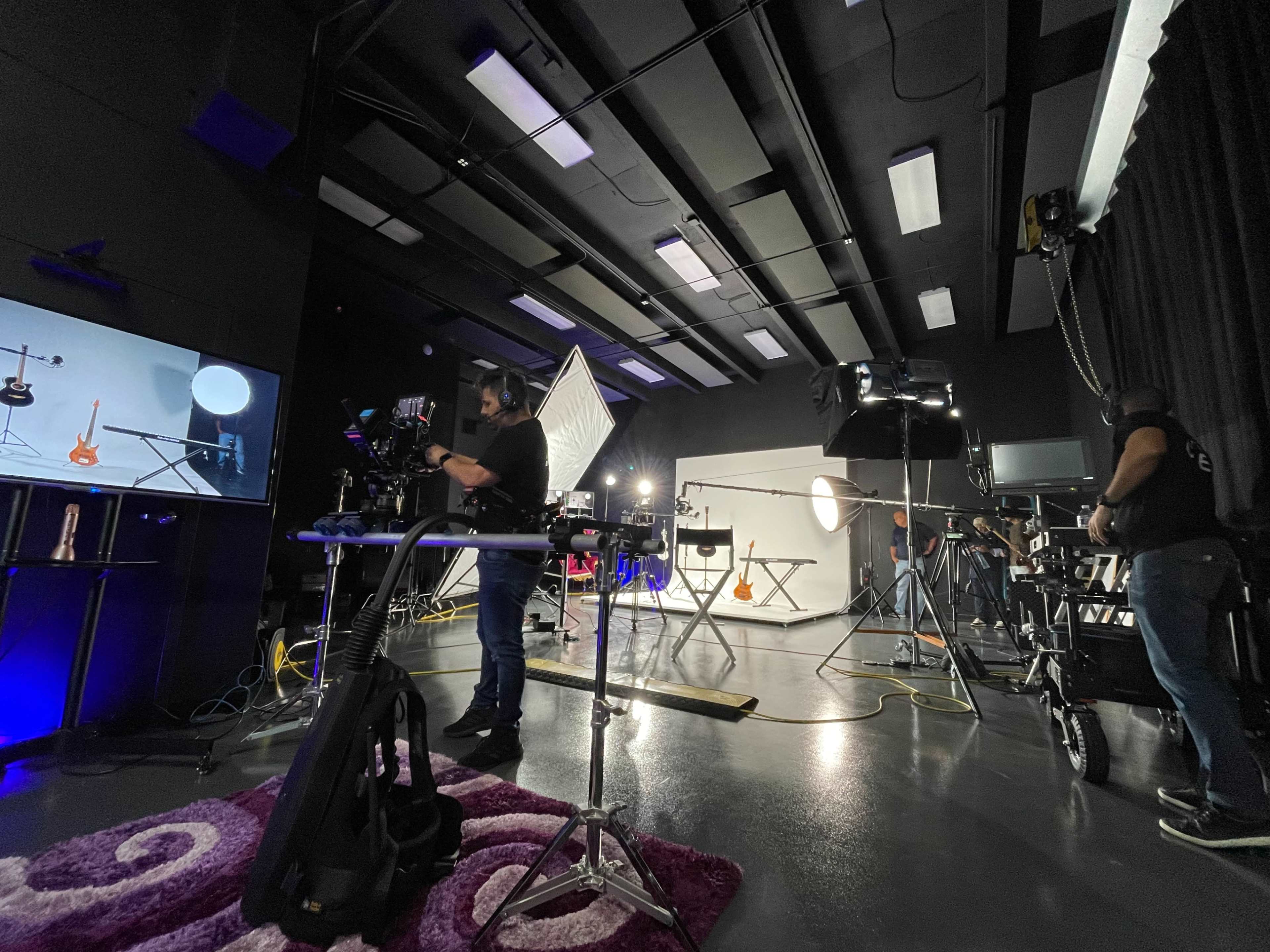 A photography studio is set up with various lighting equipment, a camera person preparing a shot, and a display screen showing a guitar setup.