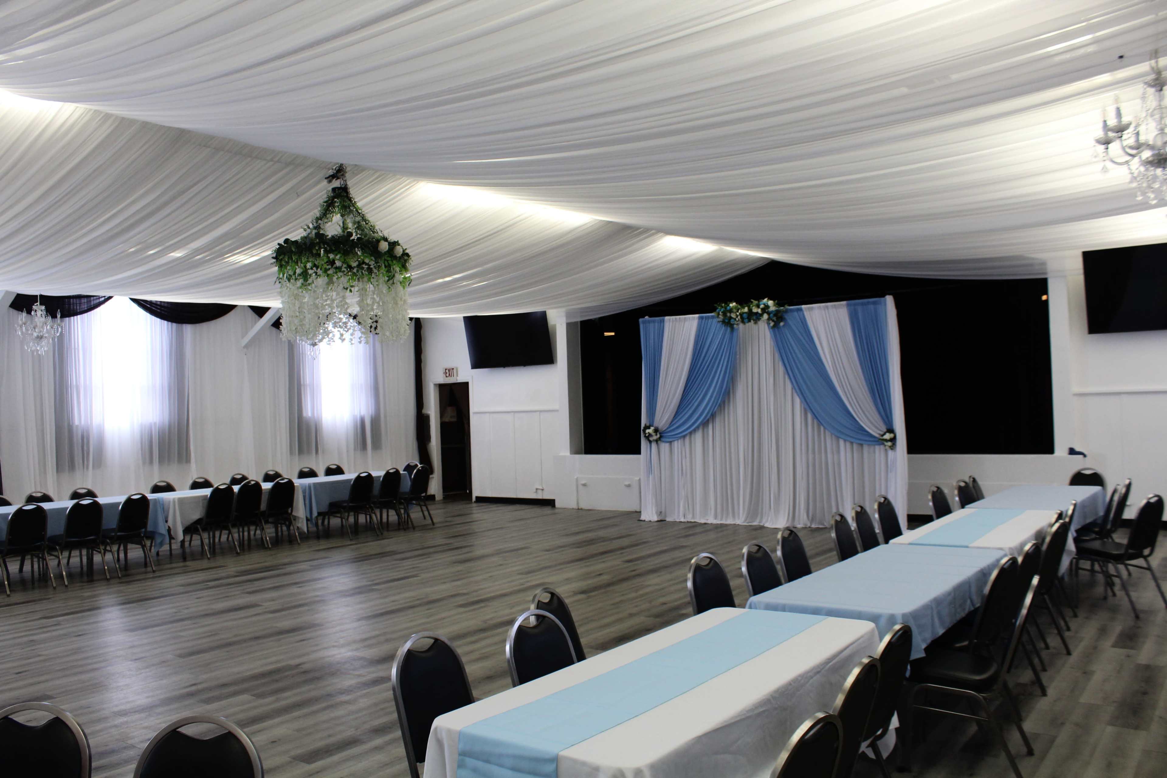 The image shows a spacious event hall featuring decorated tables with light blue tablecloths and a backdrop of draped fabric and floral accents.