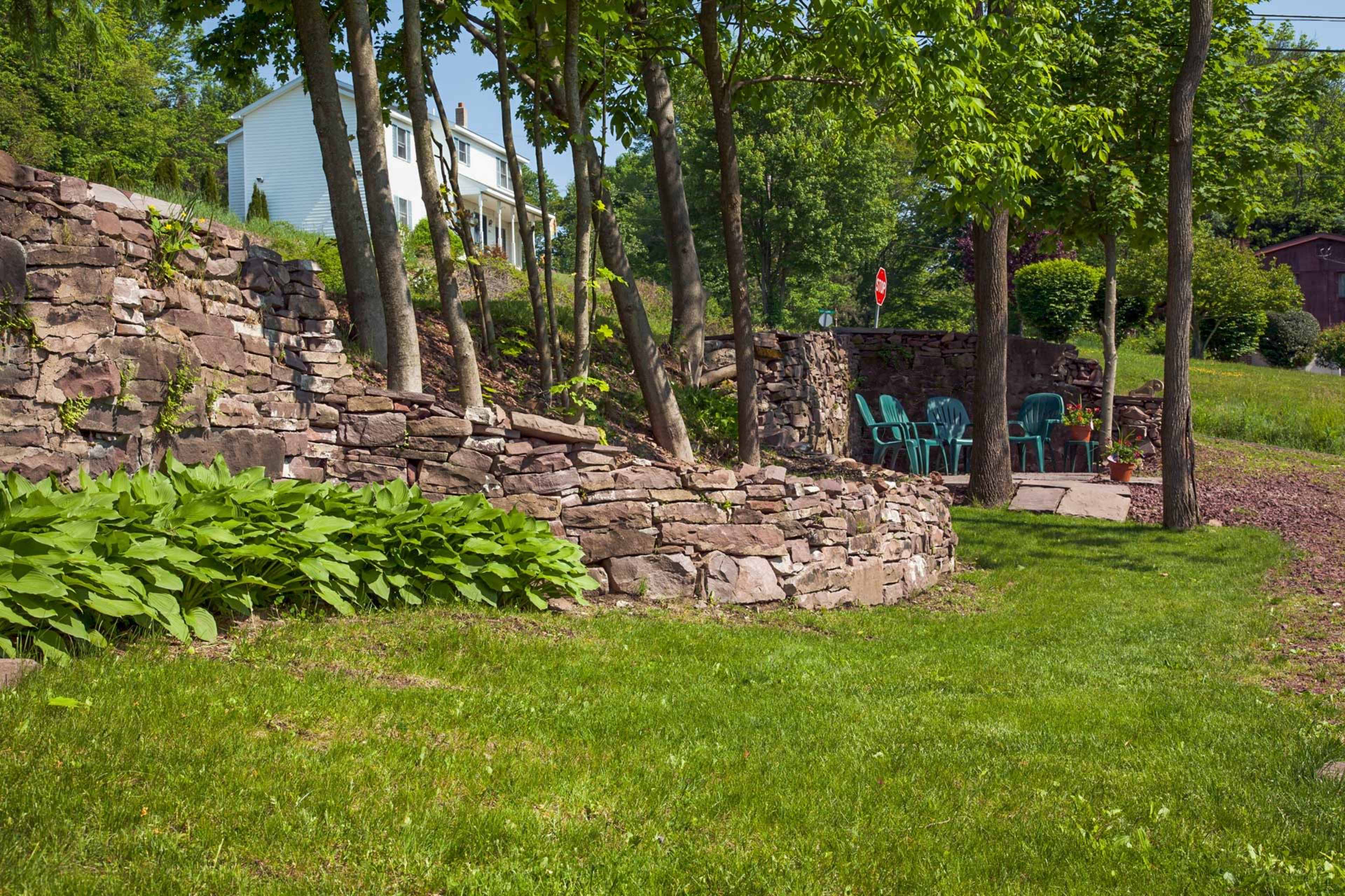 The image shows a landscaped yard with stone walls, green grass, and a pair of blue chairs under shaded trees.