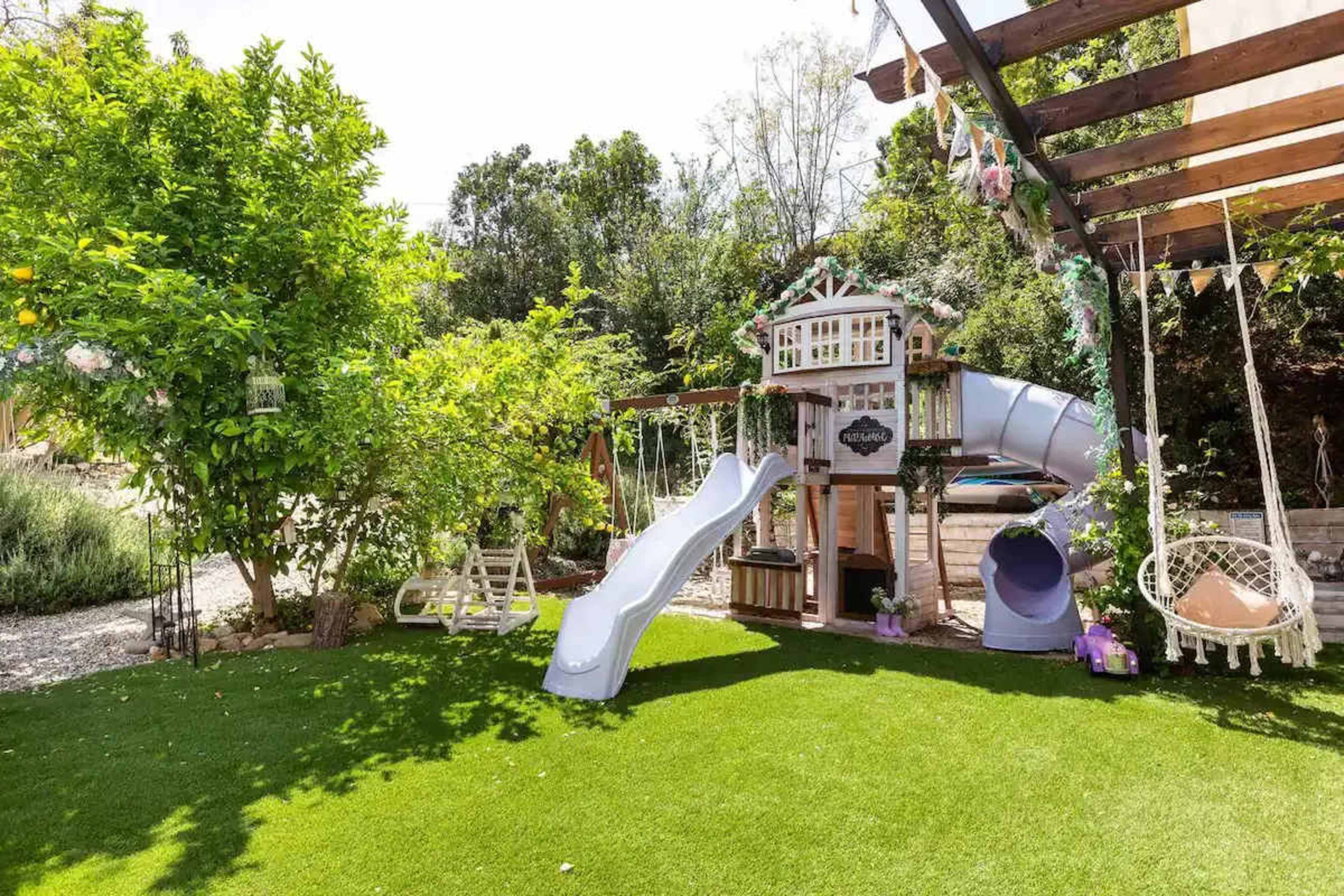 The image shows a children's play area with a wooden playhouse featuring a slide, swing set, and seating on a grassy surface surrounded by trees.