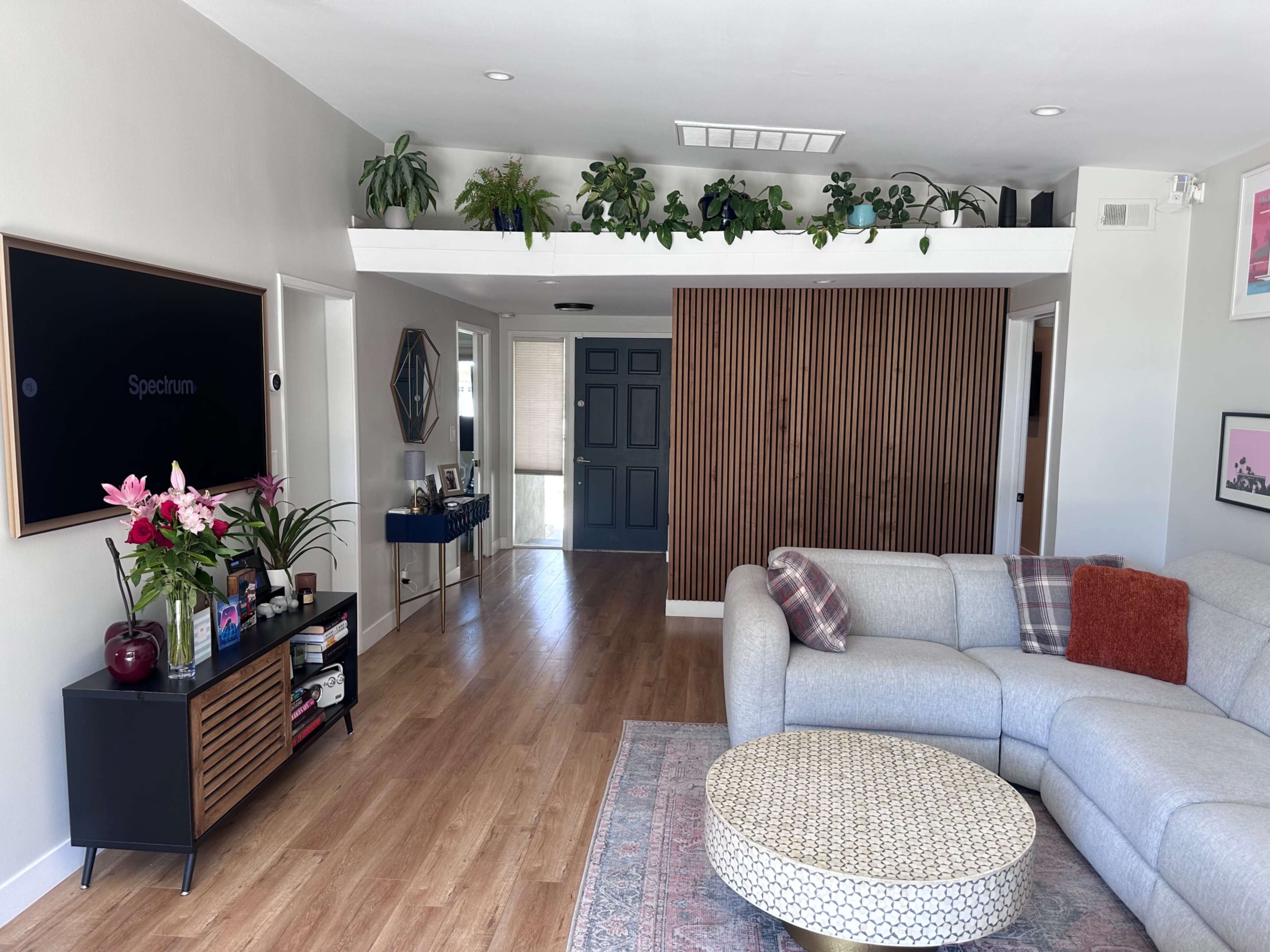 The image shows a modern living room featuring a light gray sectional sofa, a coffee table, and a wall with wooden paneling, alongside plants and decorative elements.