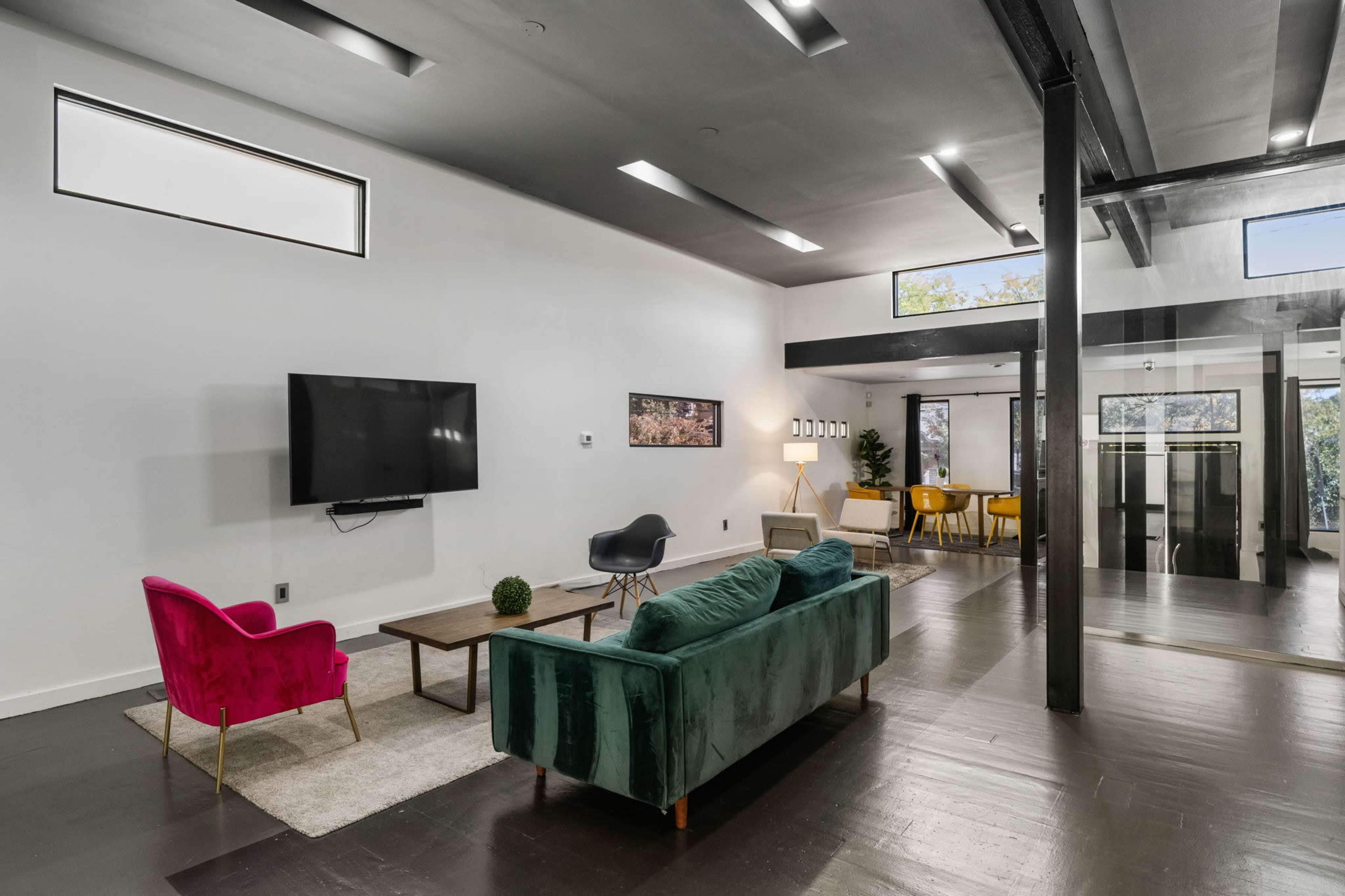 A modern living area with a pink chair, a green sofa, a coffee table, a large television mounted on the wall, and a glass-walled office space in the background.
