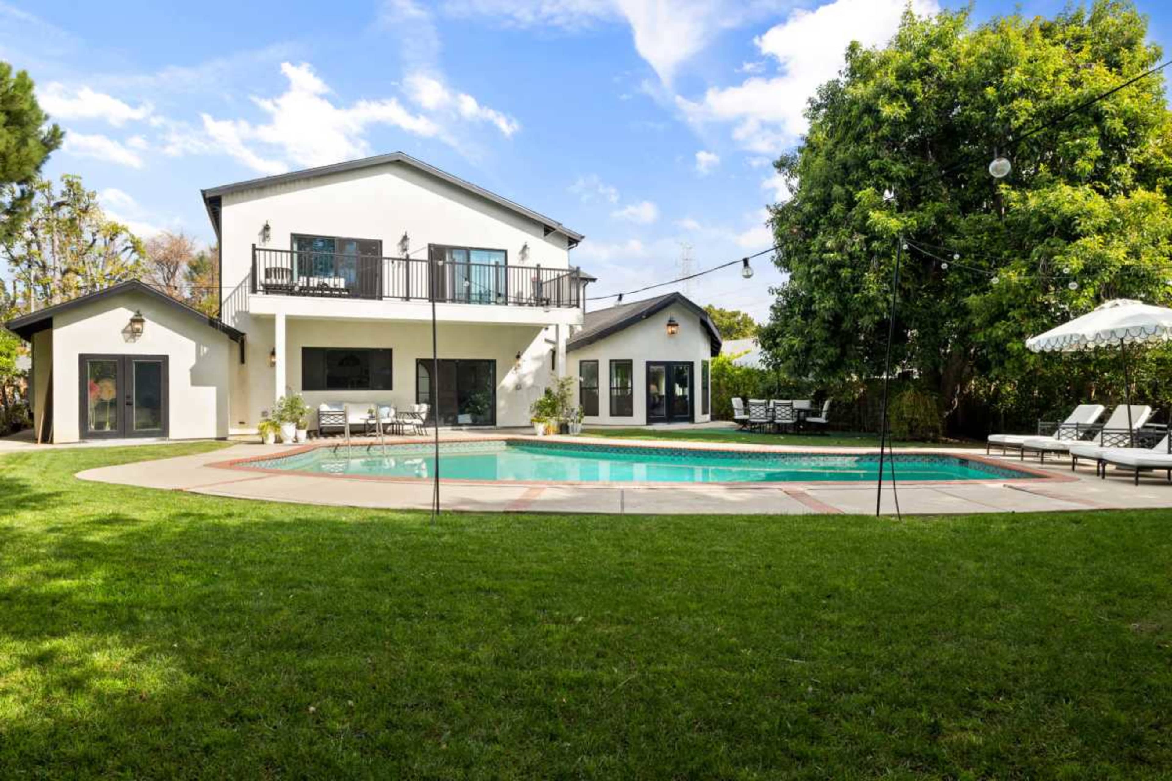 A modern two-story house with a pool and patio is surrounded by a grassy yard and trees.