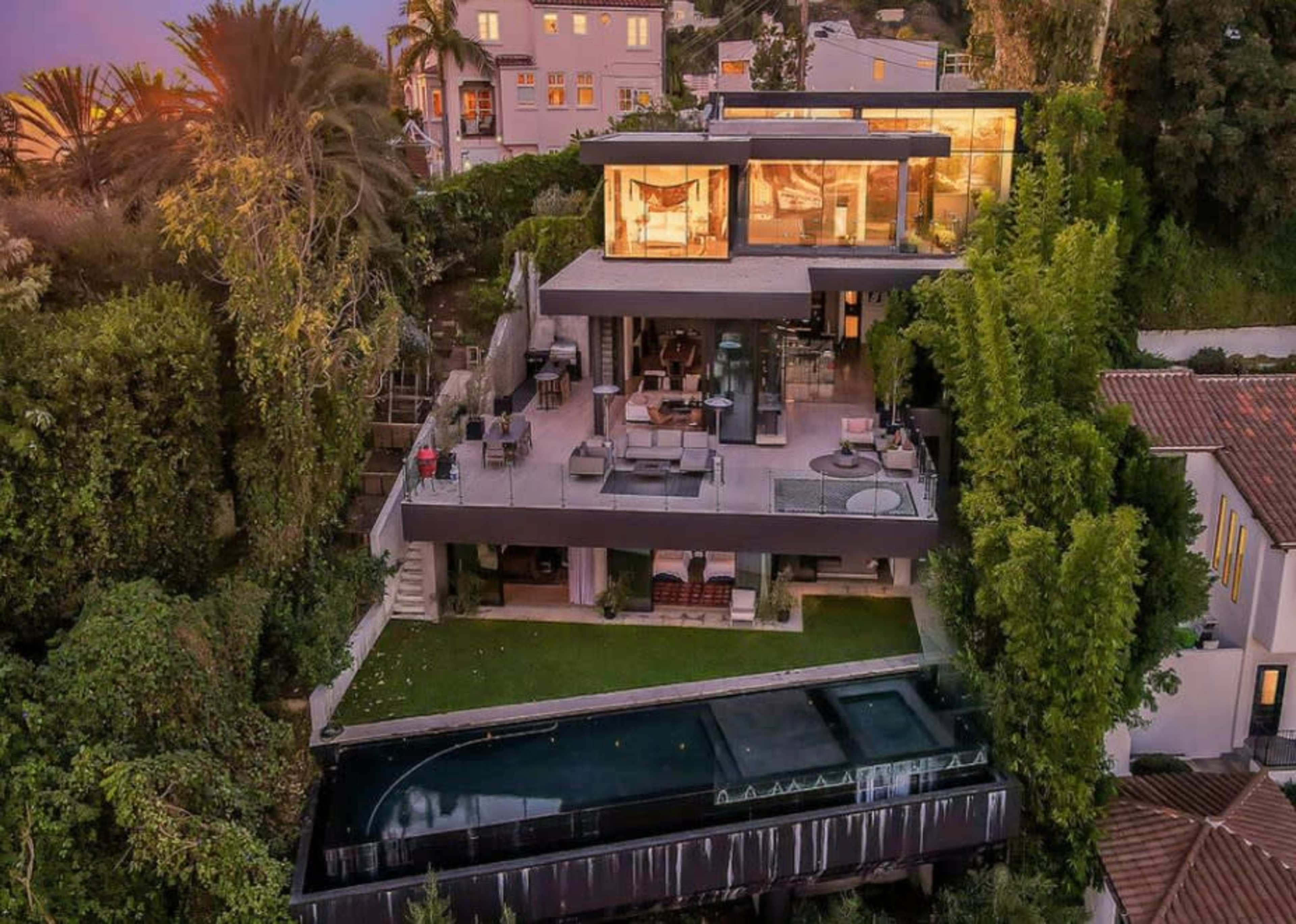 The image shows a modern three-story house built into a hillside, featuring large glass windows, multiple outdoor seating areas, and a swimming pool.