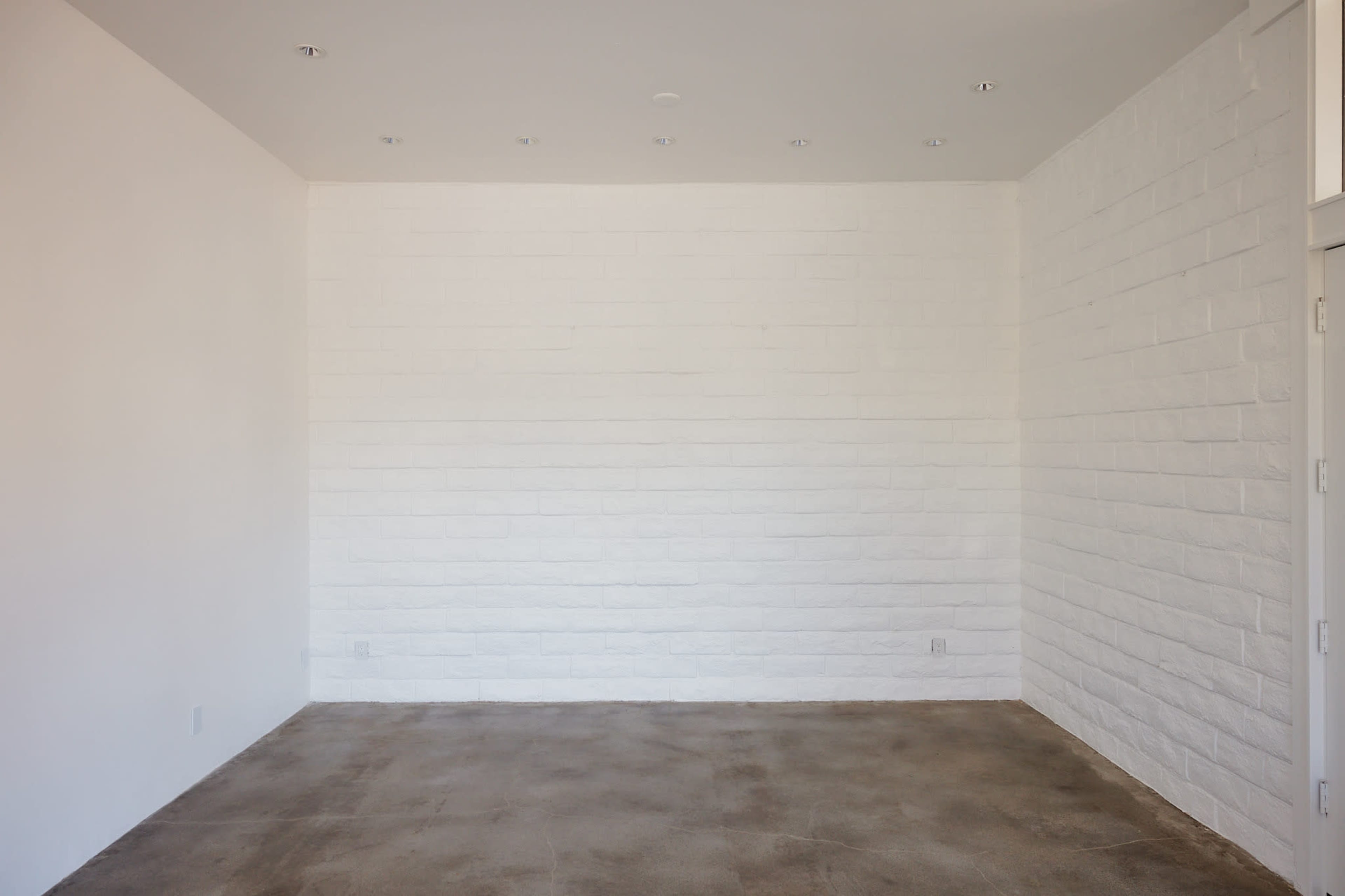 The image shows a sparsely furnished room with a white wall on one side and a concrete floor.