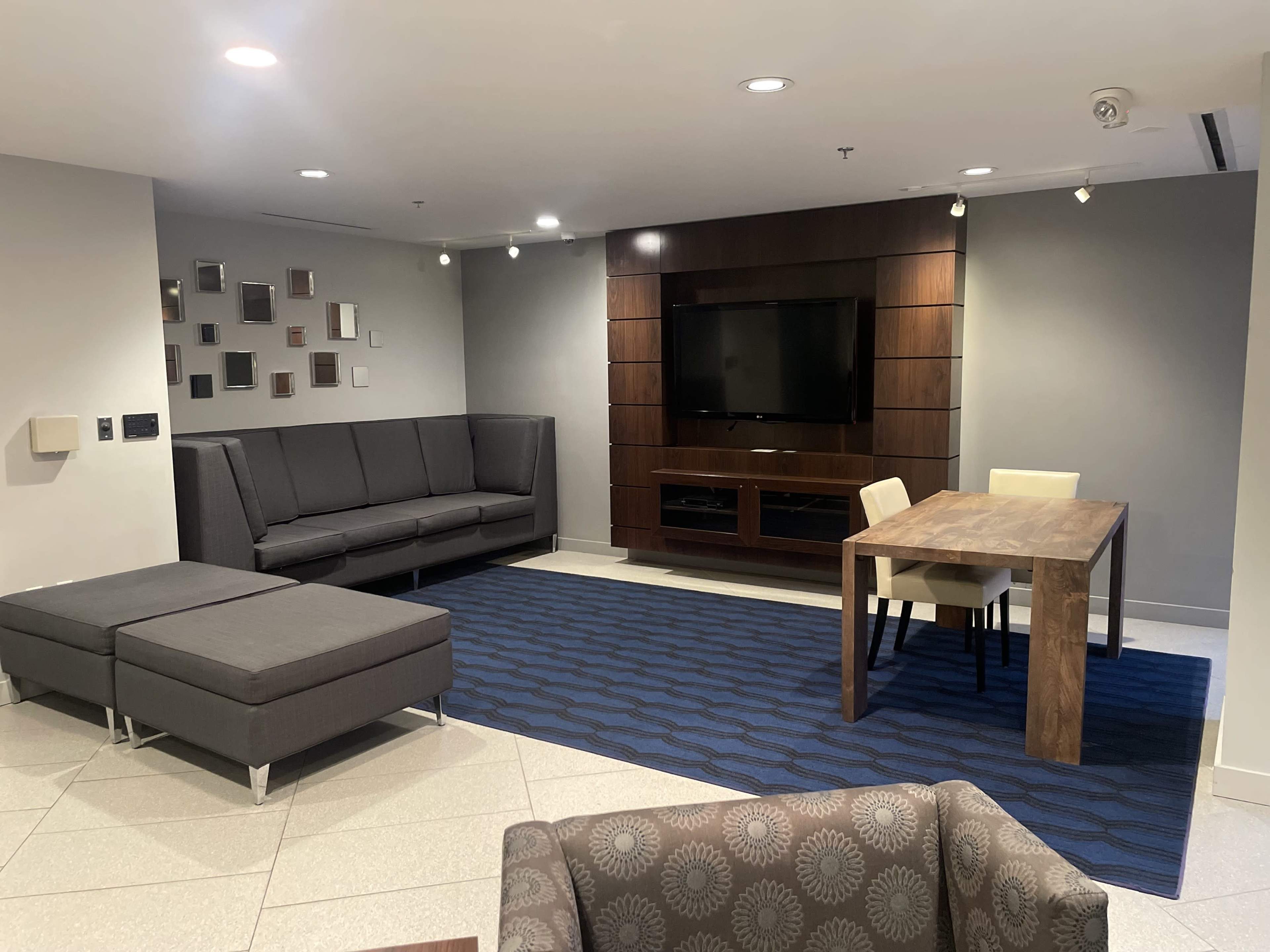 A modern lounge area with a gray sectional sofa, a wooden dining table with two chairs, and a wall-mounted television in a well-lit space.