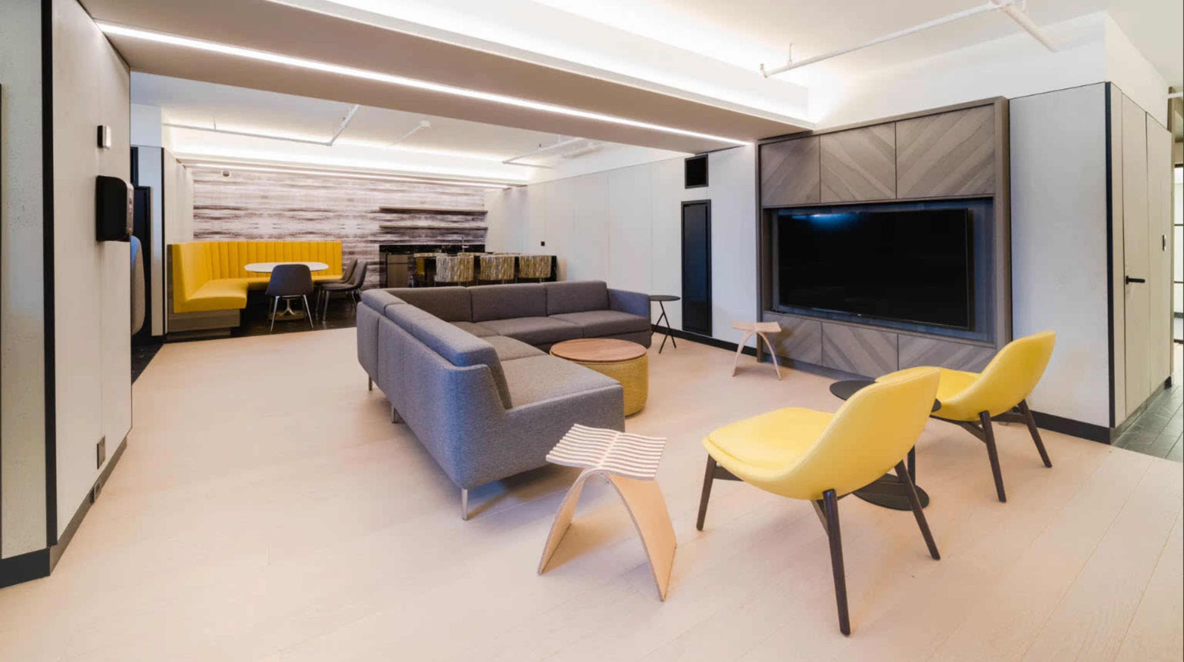 A modern, open lounge area featuring a gray sofa, yellow chairs, a round wooden coffee table, and a large television mounted on the wall.