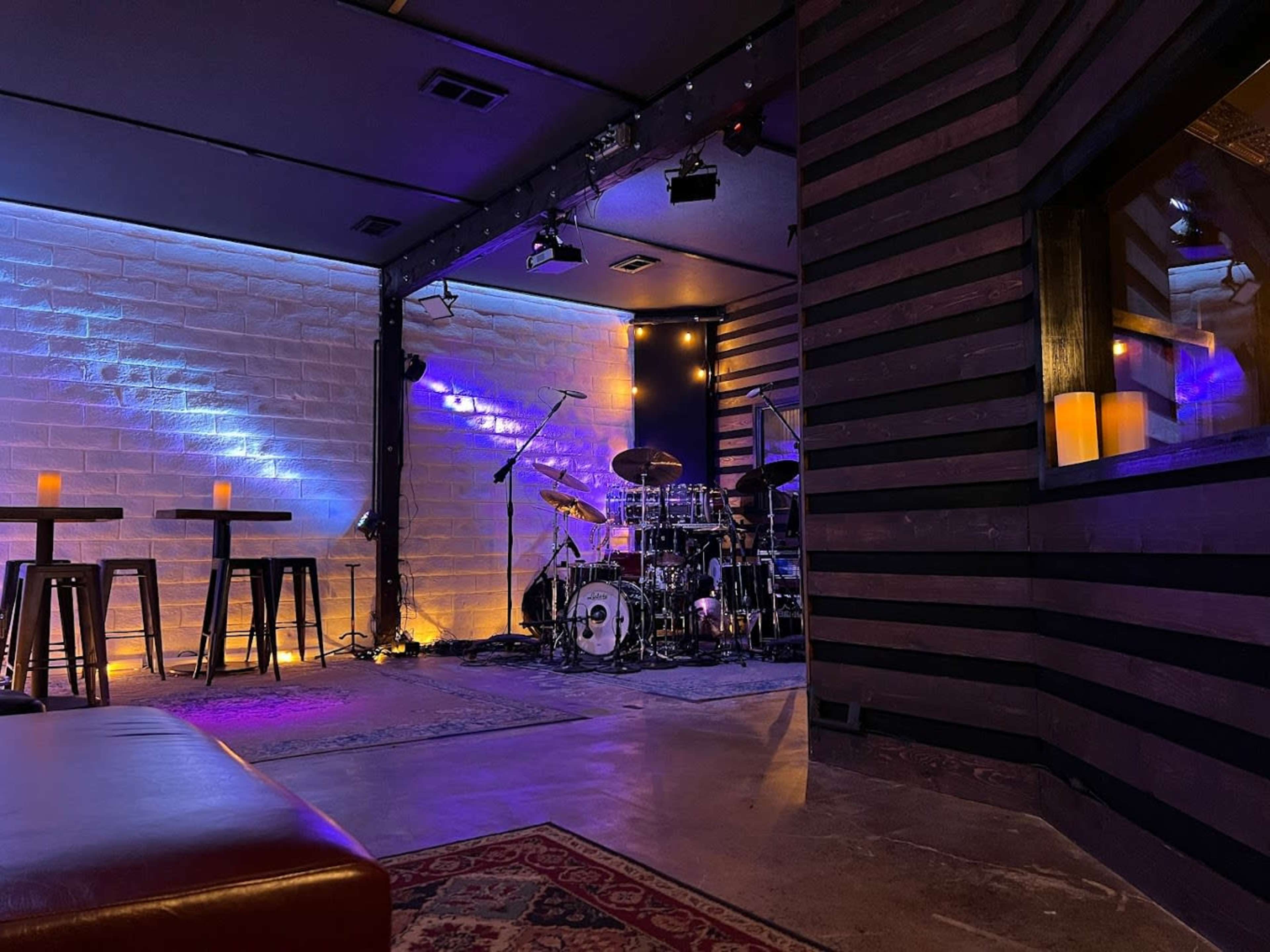 A dimly lit music venue with a stage set up for a band, featuring a drum kit and ambient lighting, alongside several tables and stools.