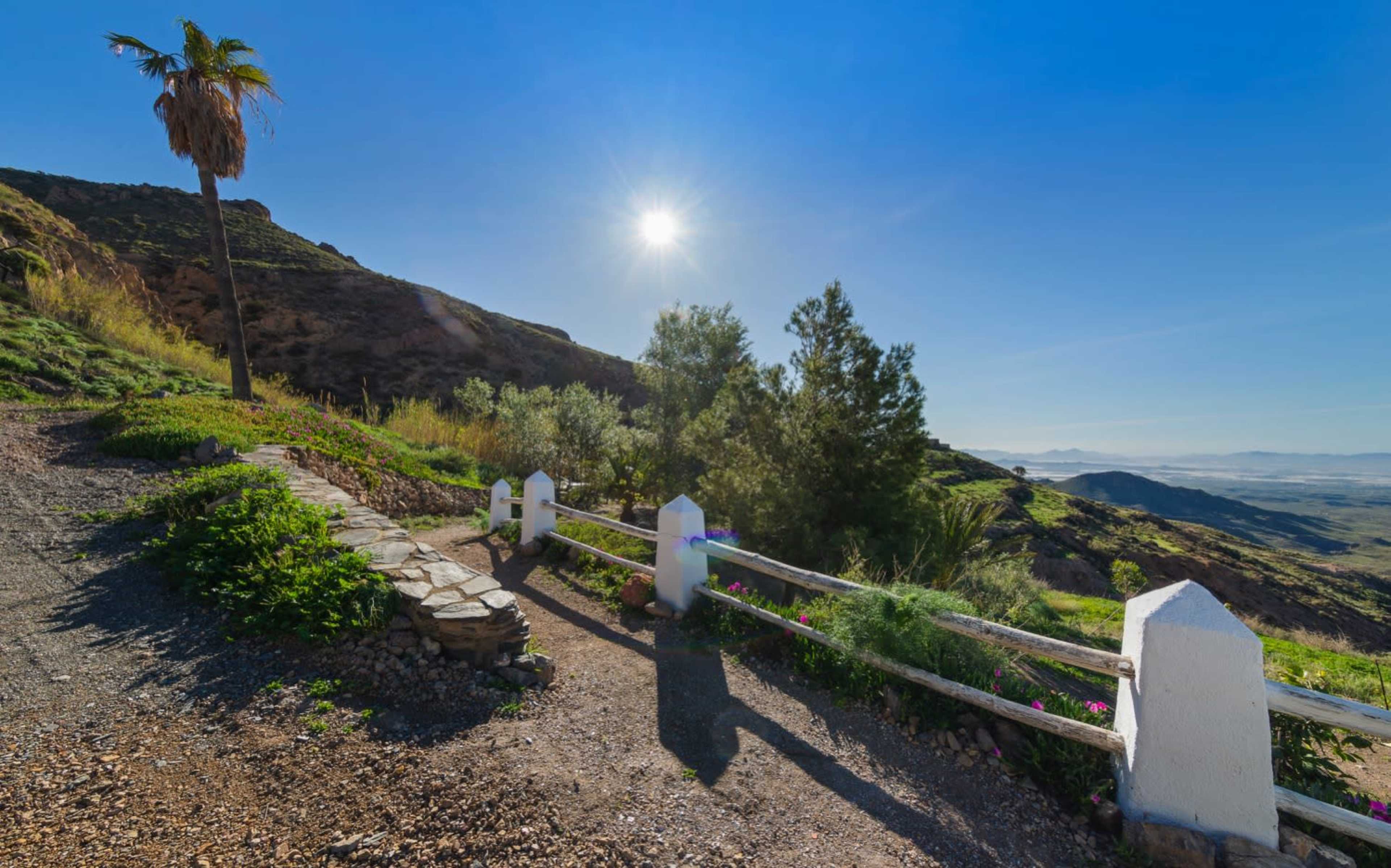 A dirt path lined with a white fence leads through green vegetation towards a mountainous landscape under a clear blue sky.
