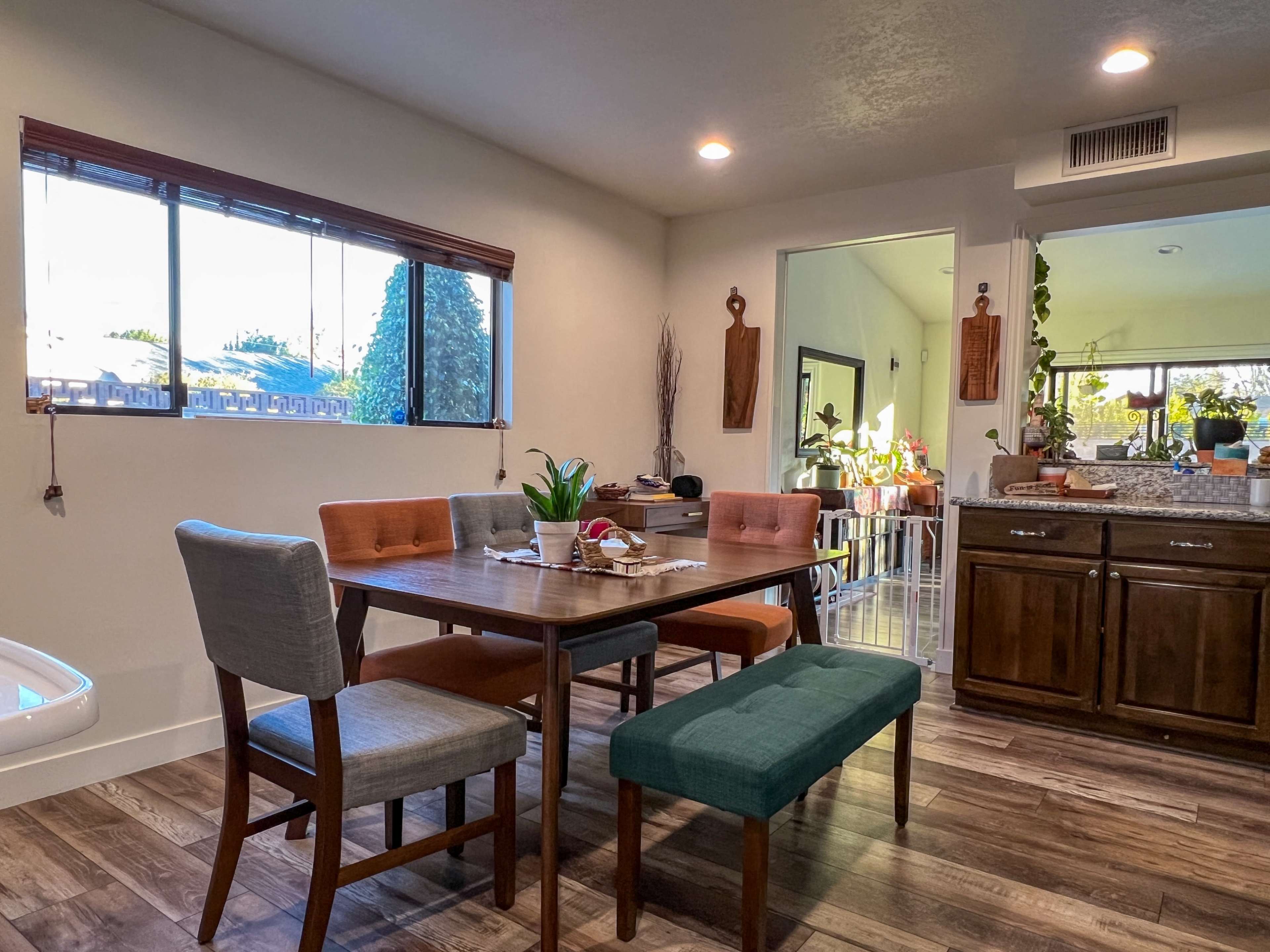 The image shows a dining area with a wooden table surrounded by chairs and a bench, adjacent to a kitchen with visible cabinets and counter space.