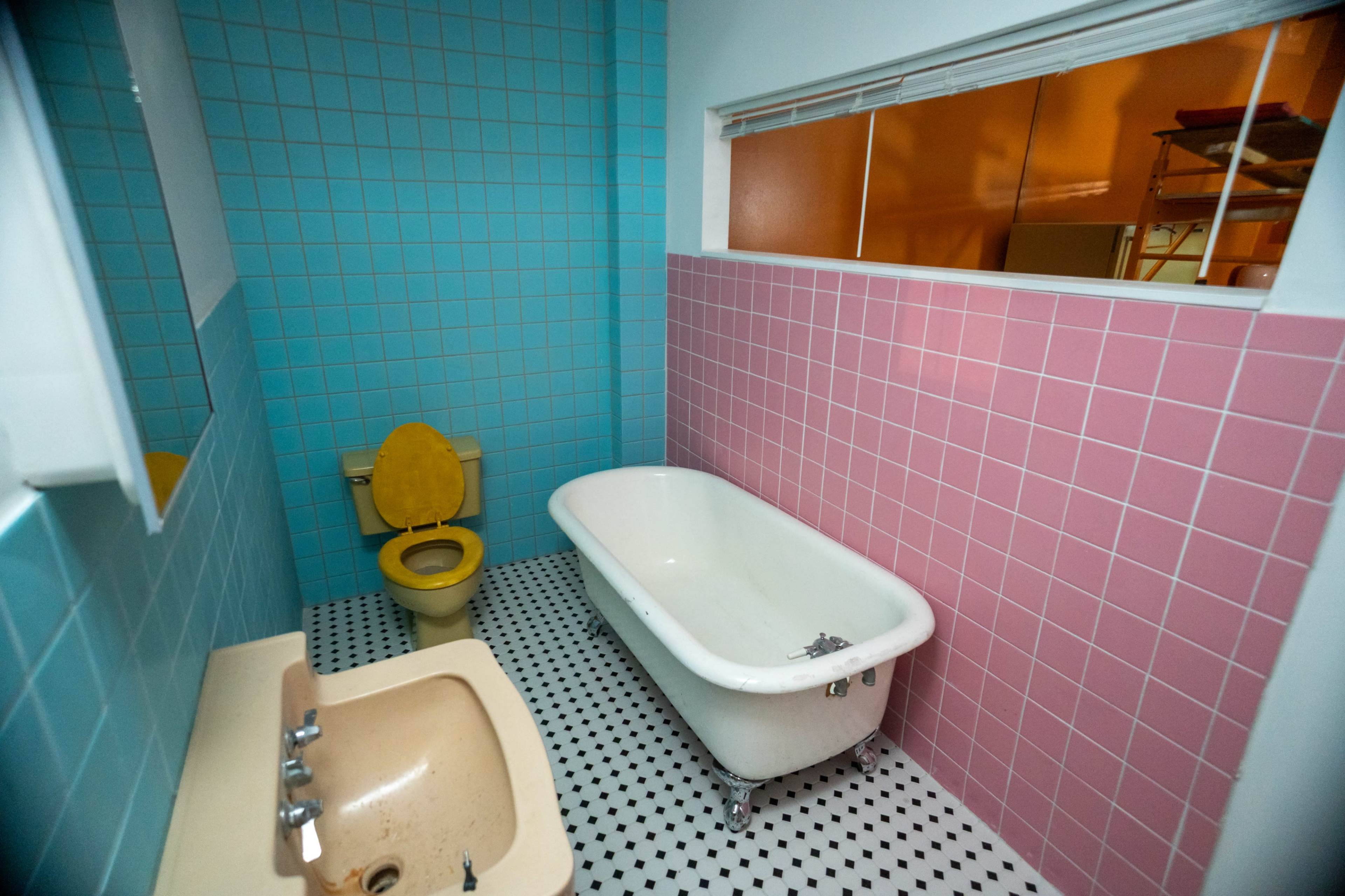 The image shows a small bathroom featuring a yellow toilet, a pink bathtub, and blue and pink tiled walls.