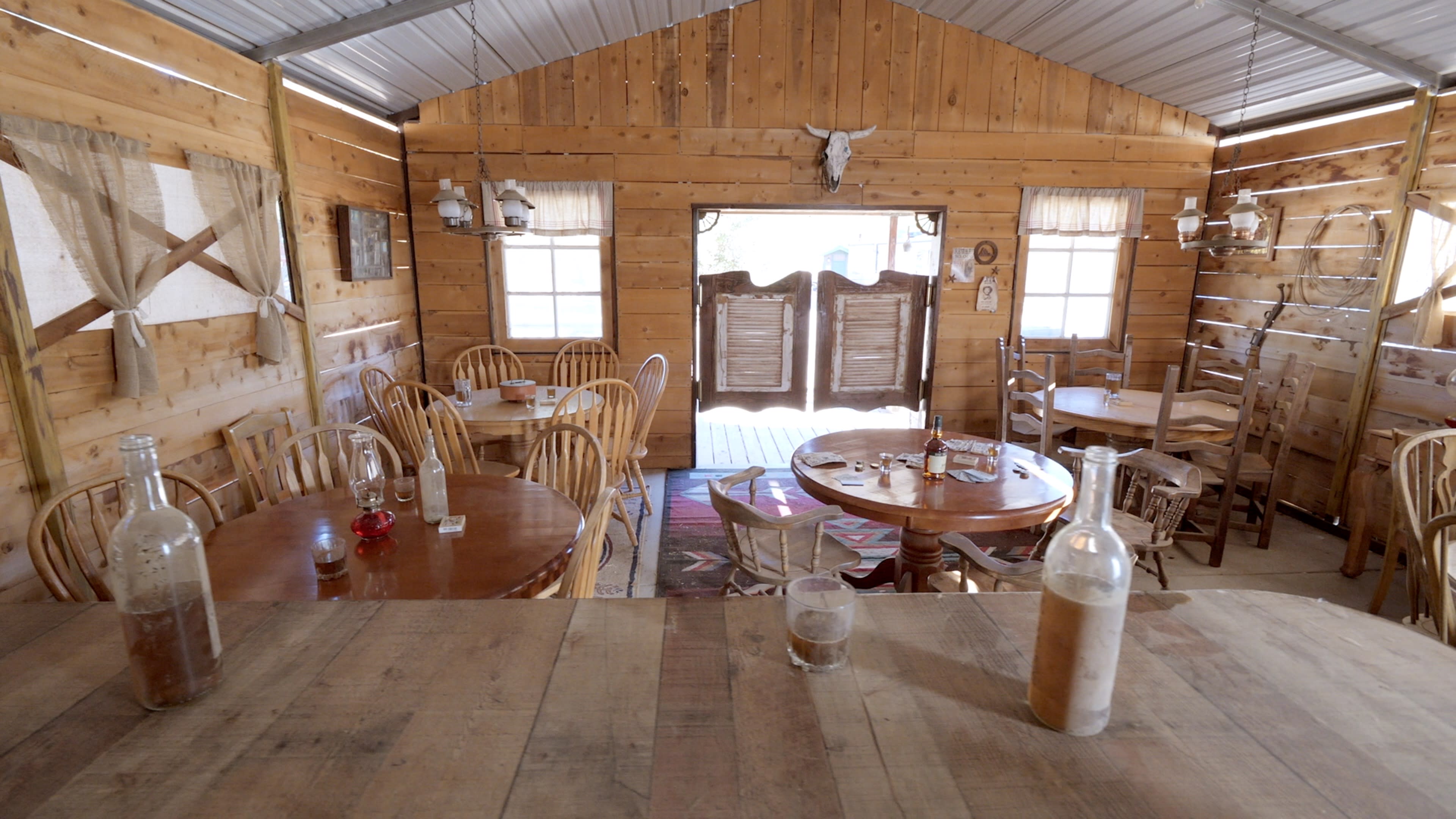 The interior of a rustic wooden restaurant features several round tables, chairs, and a bar area, with a wooden door leading outside.