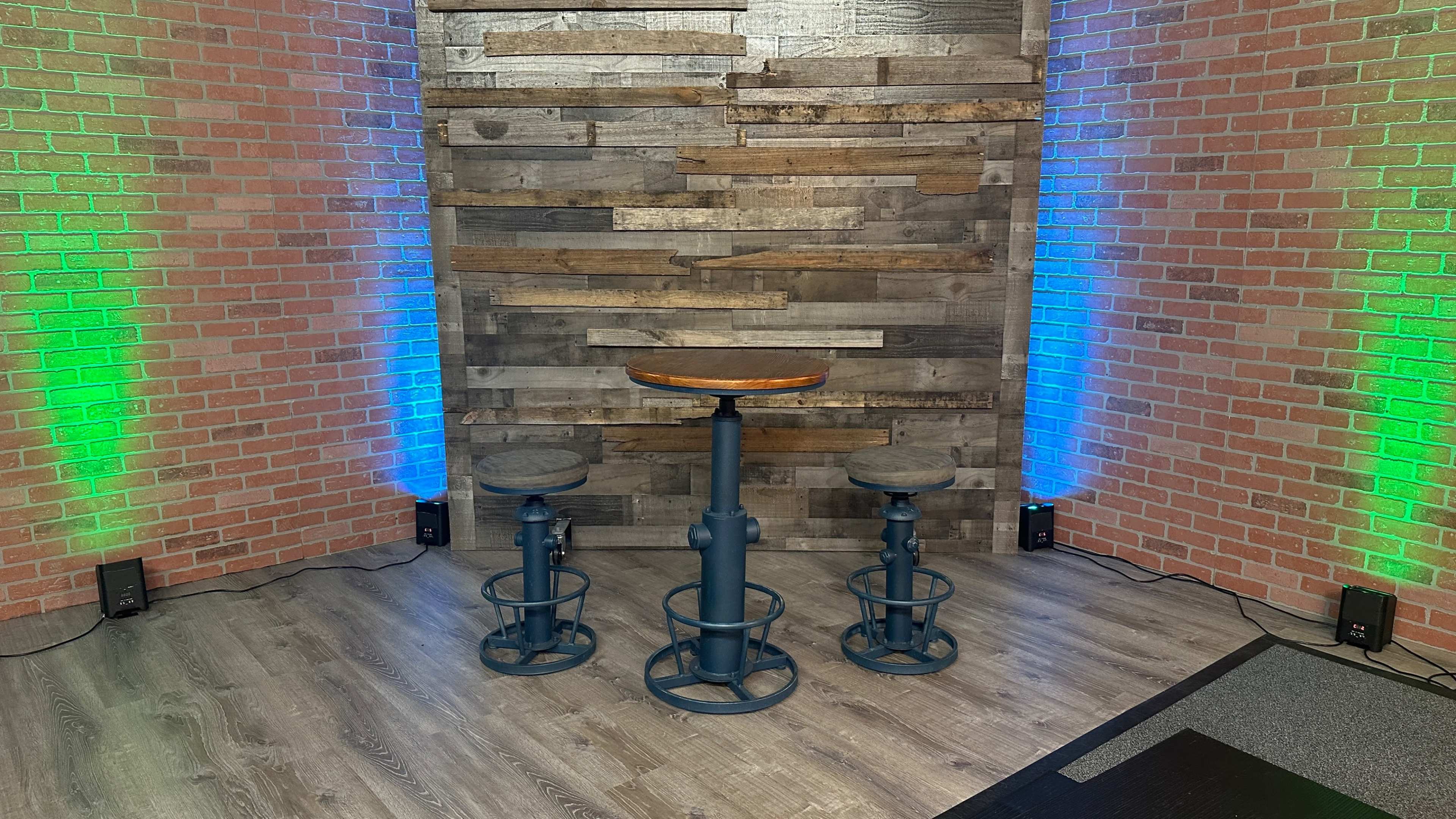 A small seating area featuring a wooden-top bar table and two stools, positioned against a textured wall made of wooden panels and brick, with green and blue lights illuminating the space.