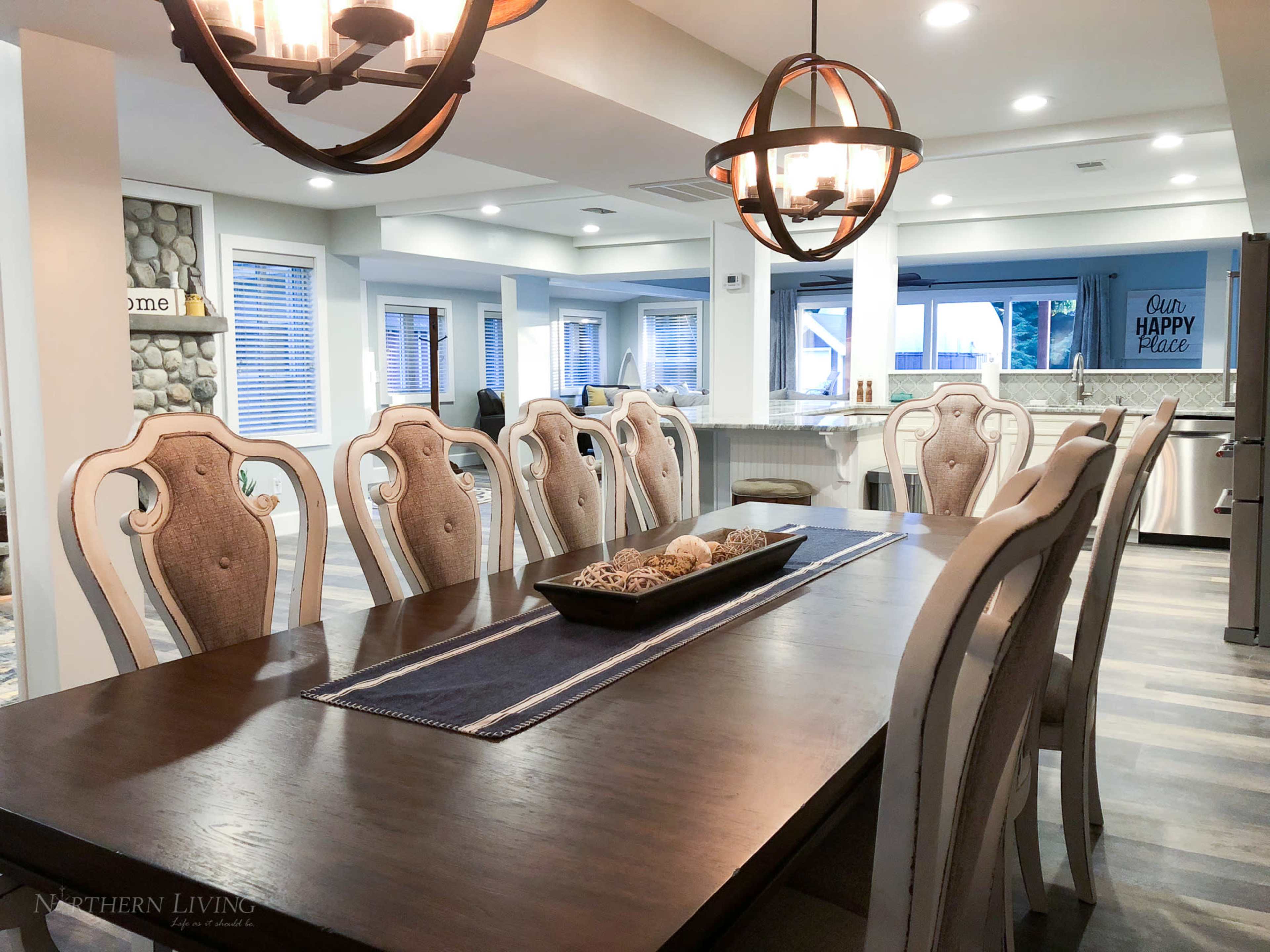 A large dining table with upholstered chairs is set in a well-lit room featuring a modern kitchen in the background.