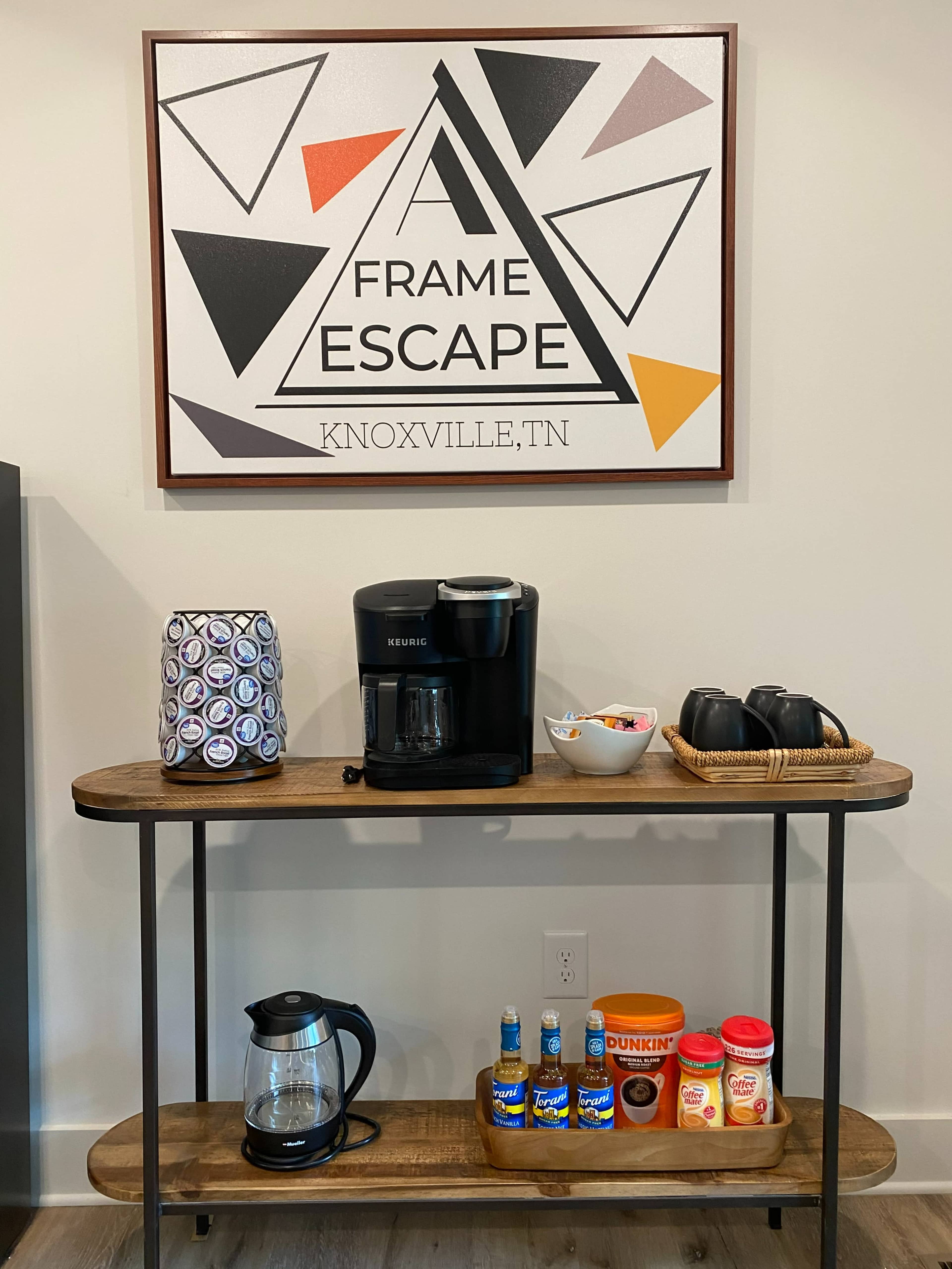 A coffee station is set up on a wooden table, featuring a Keurig machine, coffee supplies, and a decorative sign above it.