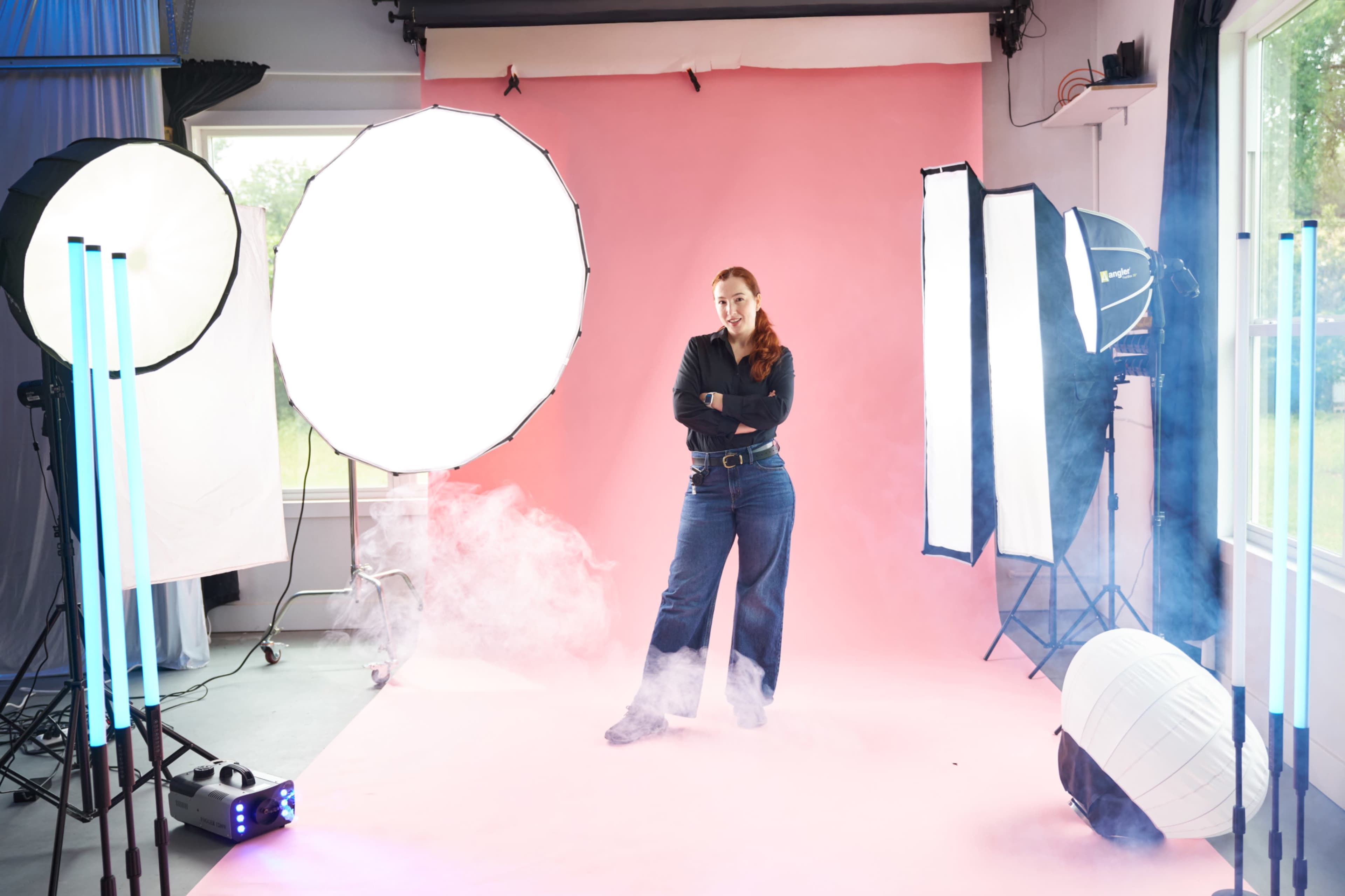 A model stands confidently on a pink backdrop in a photo studio, surrounded by various lighting equipment and fog.