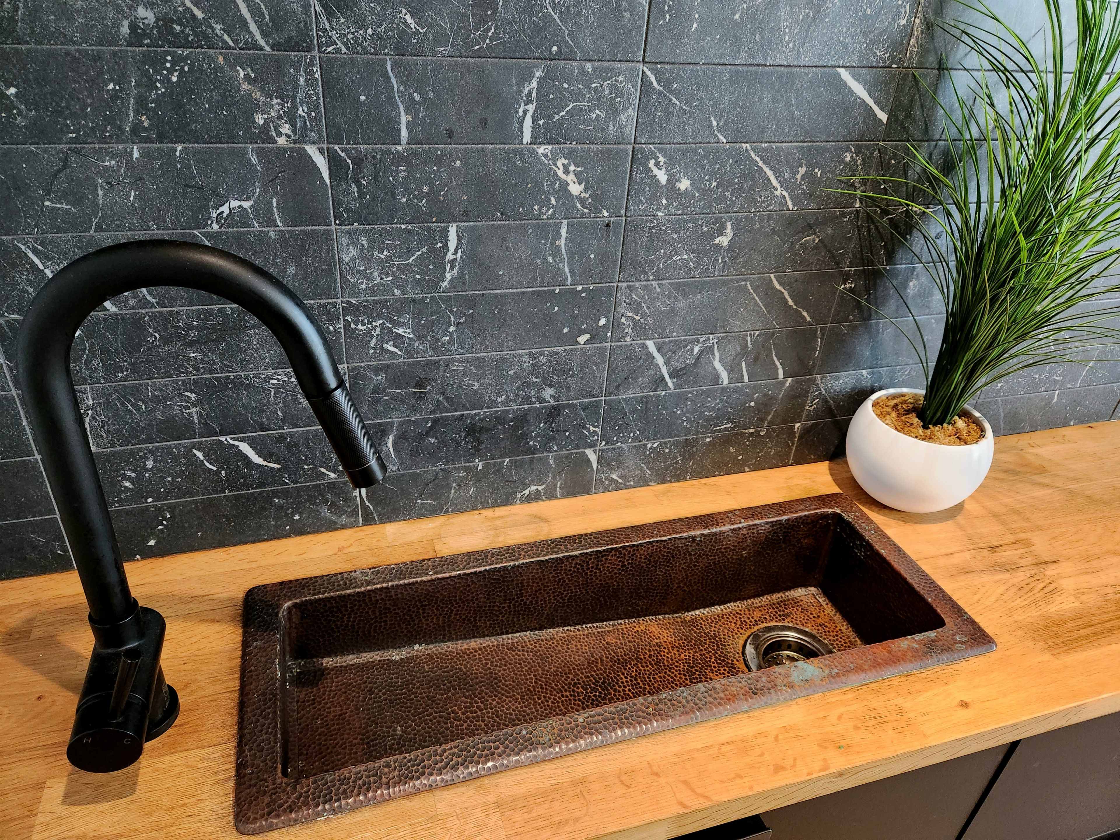 The image shows a black faucet above a copper sink set into a wooden countertop, with a potted plant in a white container on the side.