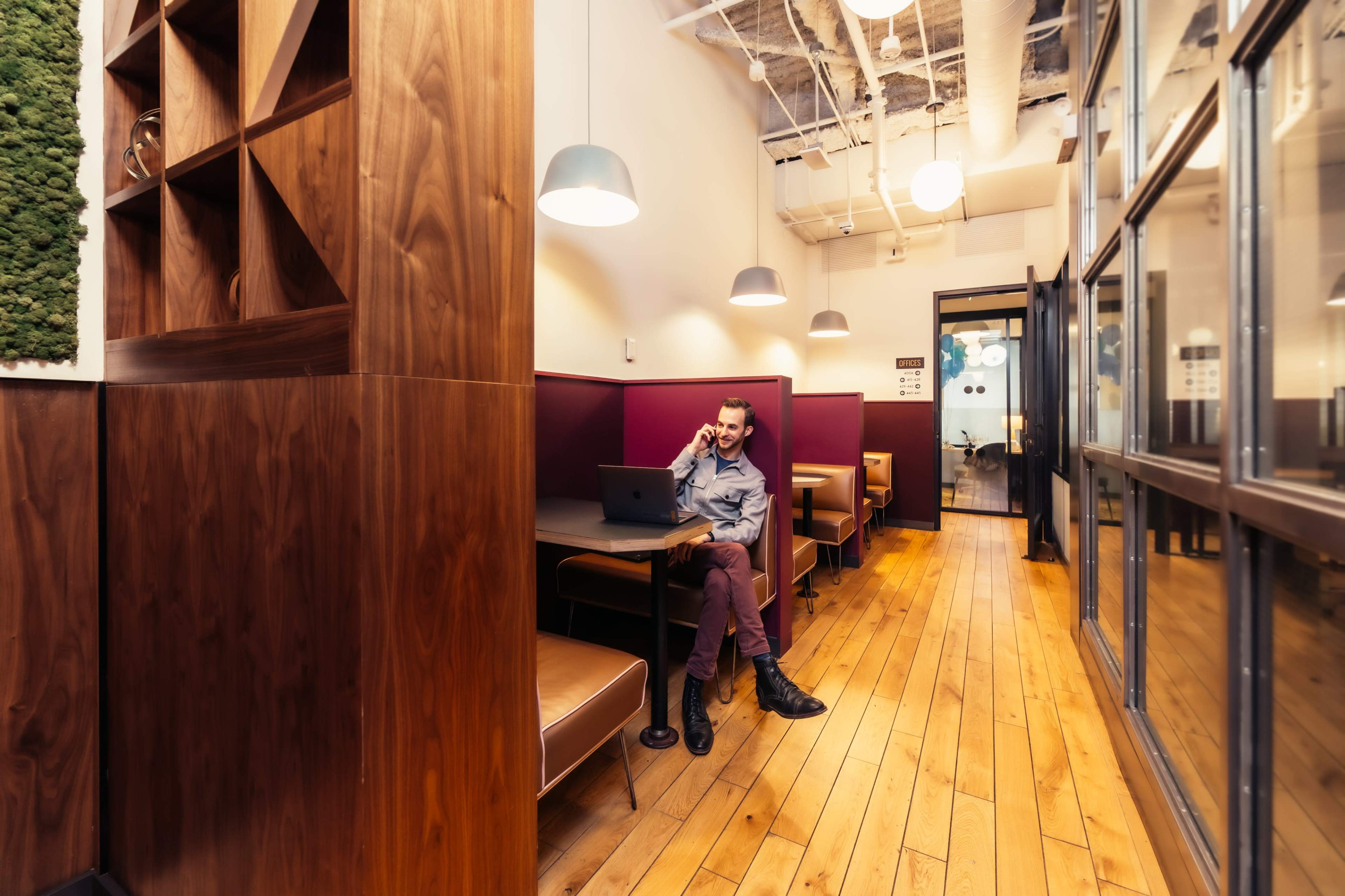 A man works on a laptop at a table in a modern office space featuring wooden floors and private seating areas.