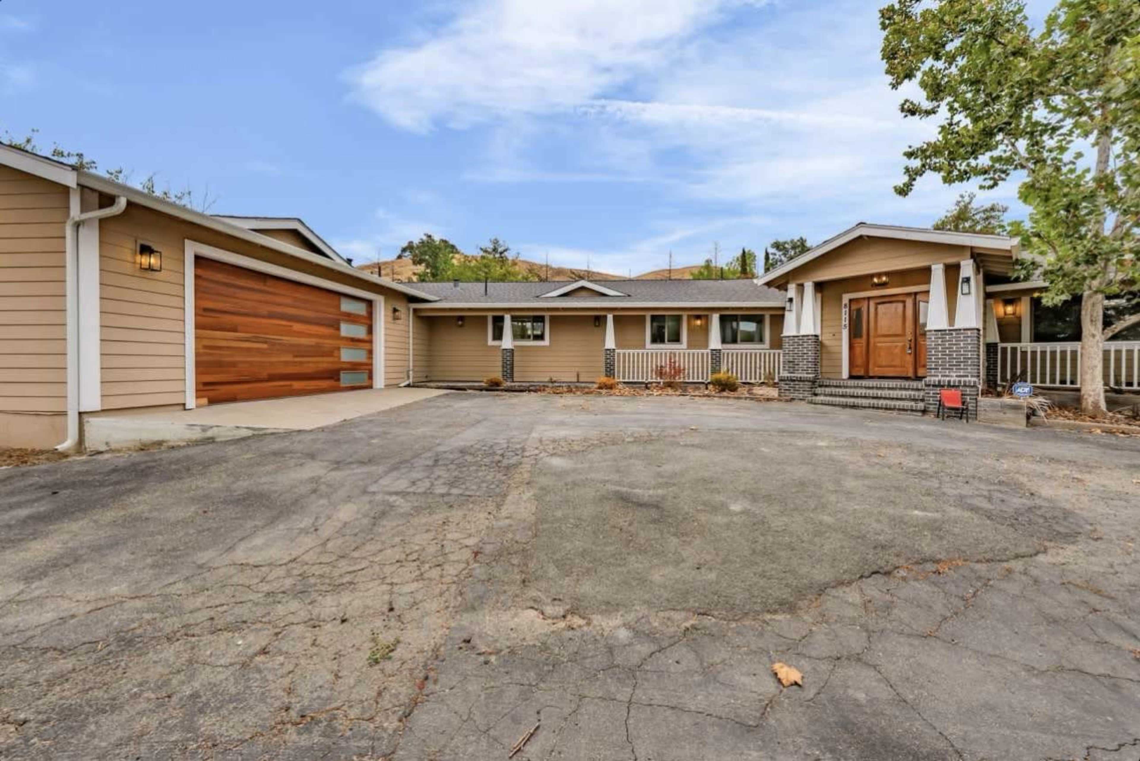 The image shows a single-story house with a textured brown exterior, a two-car garage, and a paved driveway surrounded by trees.