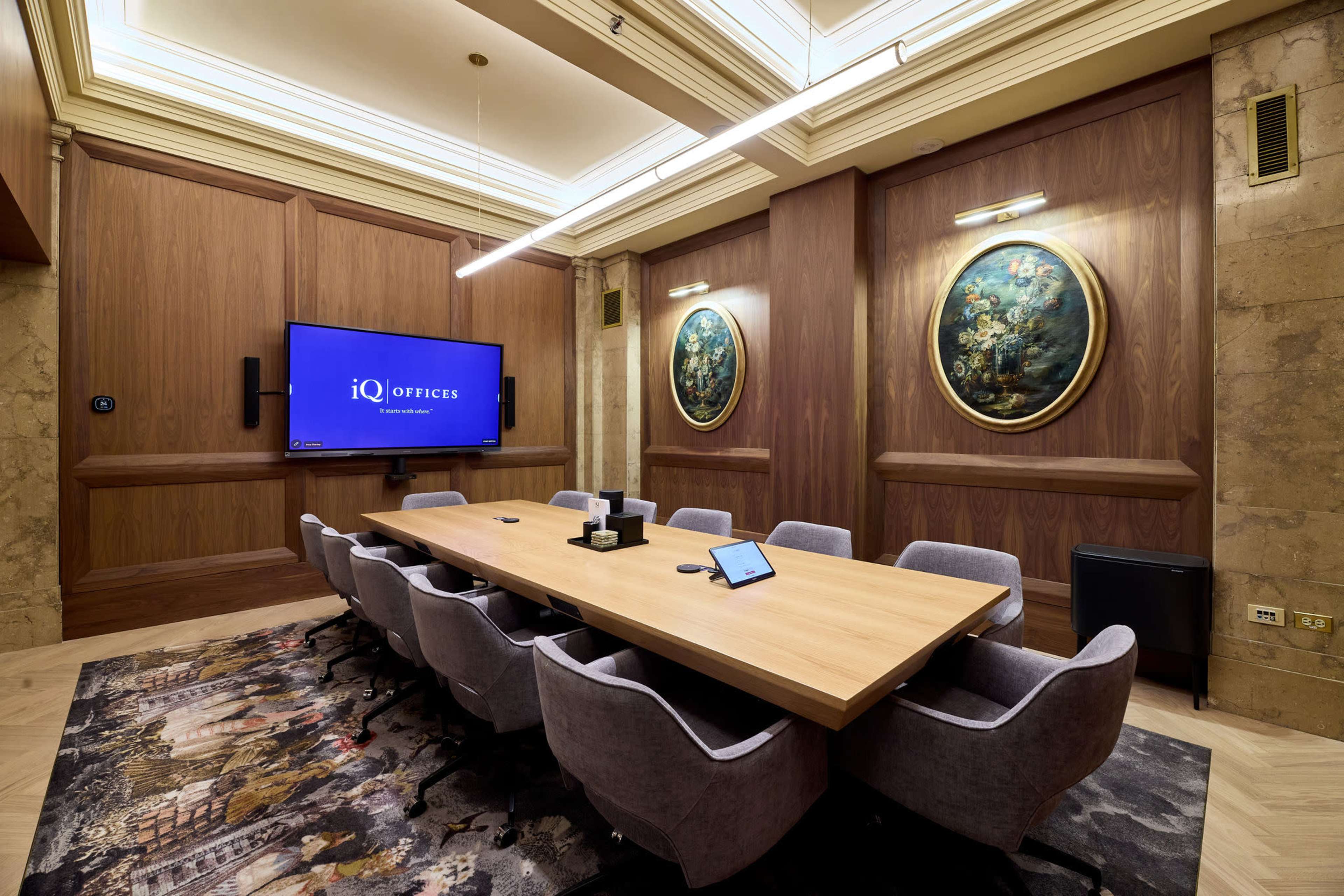 The image shows a modern conference room with a large wooden table, grey chairs, a wall-mounted screen displaying the iQ Offices logo, and framed artwork on the walls.