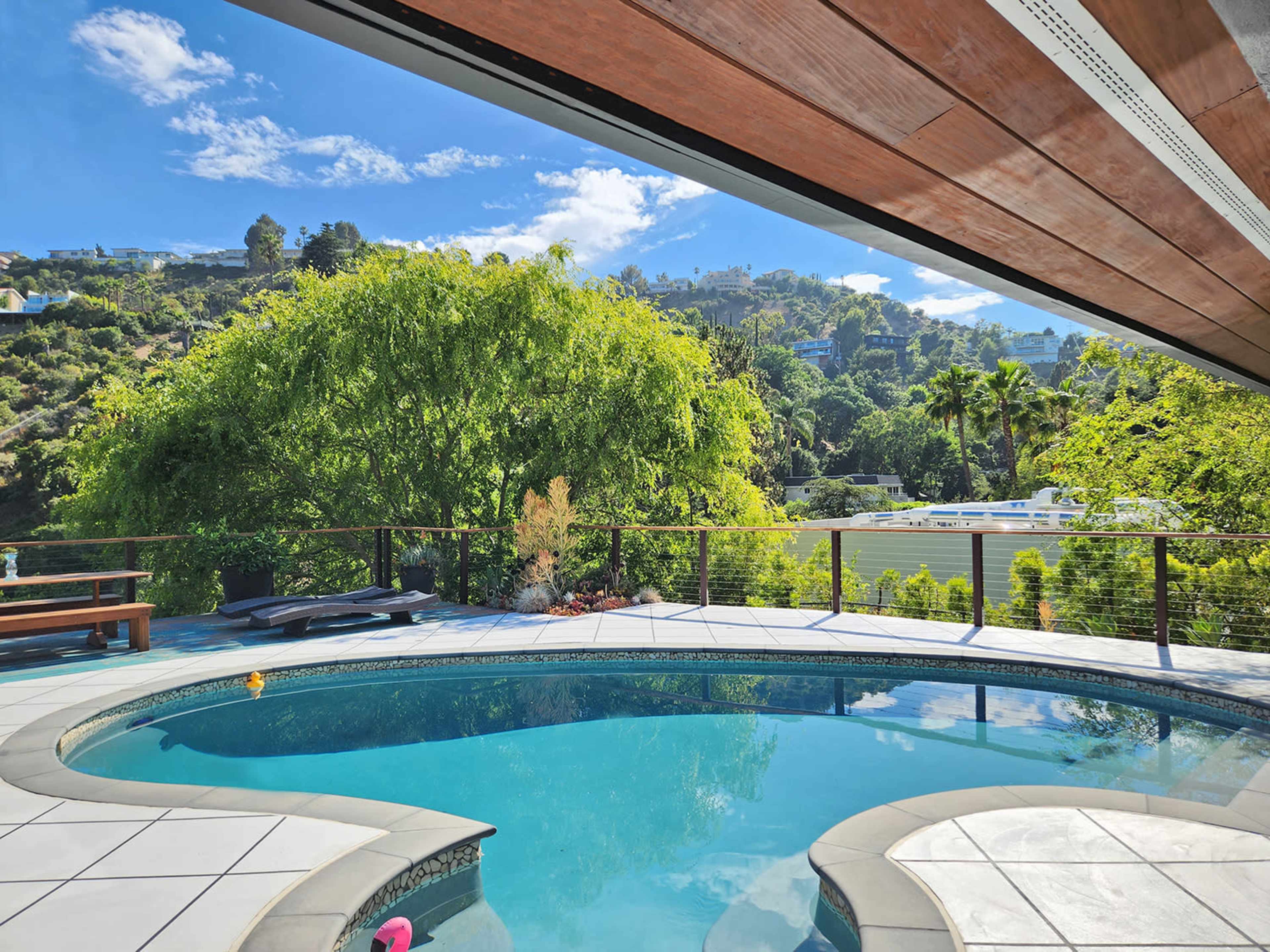 A swimming pool with a curved edge is surrounded by a deck and overlooks a lush hillside with greenery and palm trees.