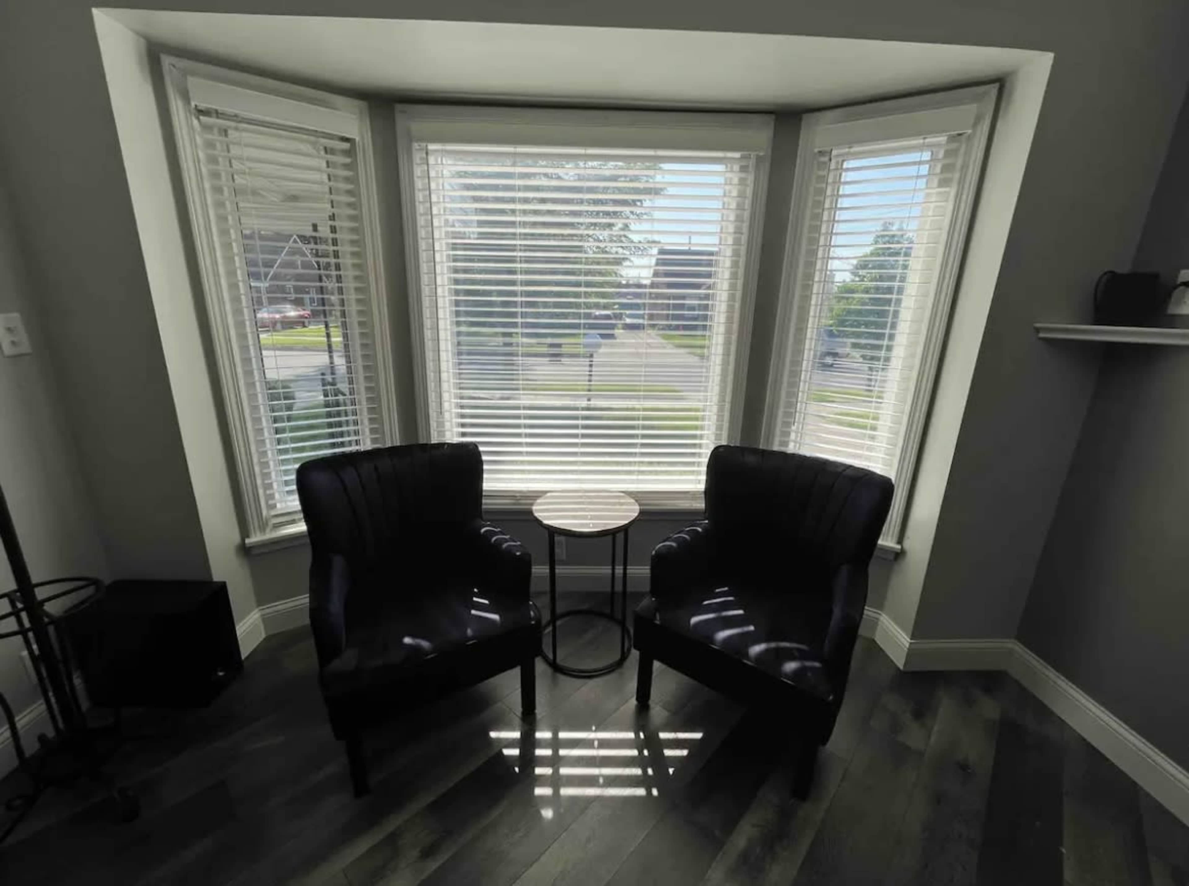 A bright corner nook with two black chairs facing a small round table, surrounded by large windows with blinds.