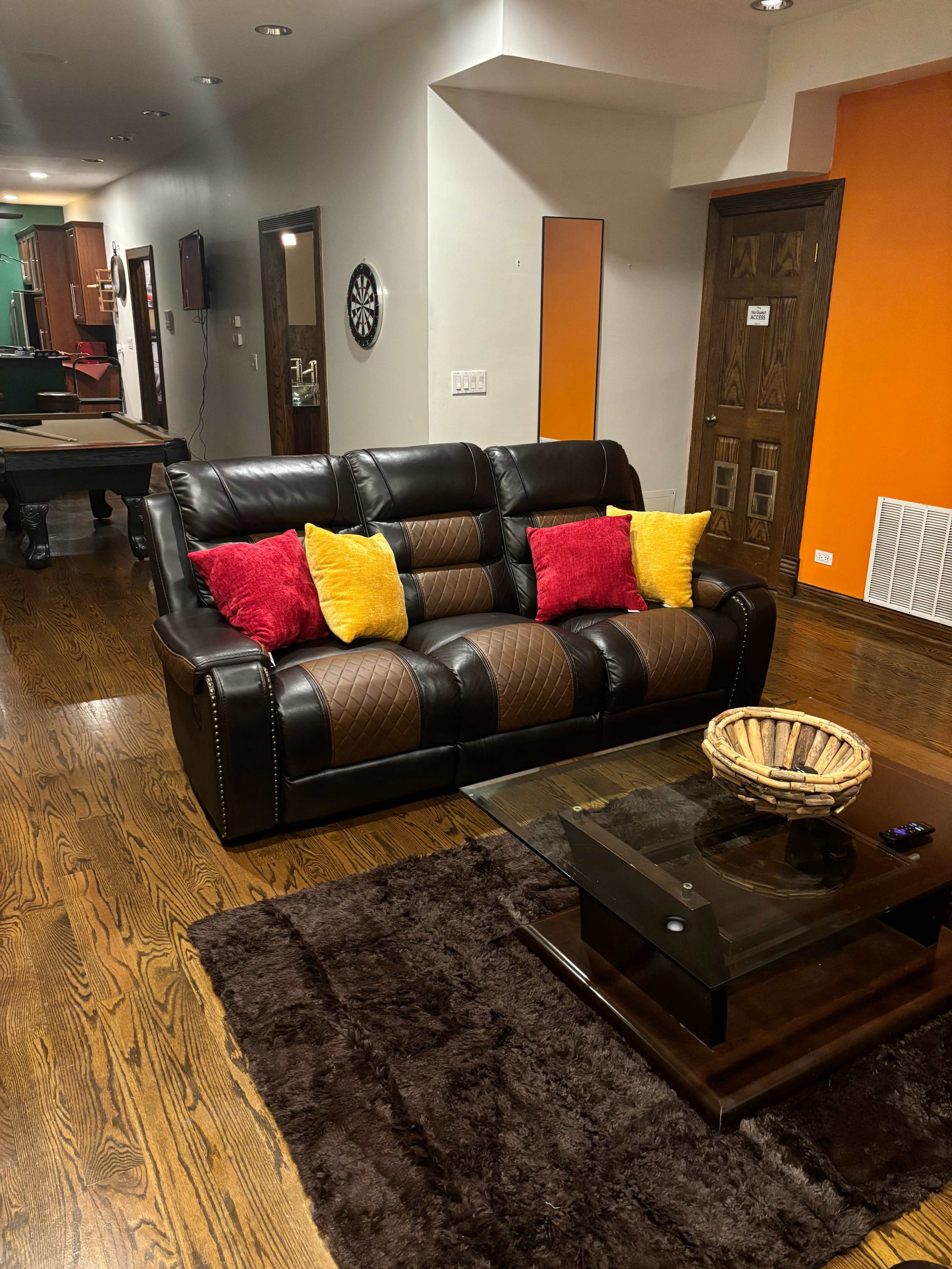 The image shows a living room featuring a dark leather couch with colorful throw pillows, a glass coffee table, and a plush brown rug on wooden flooring.