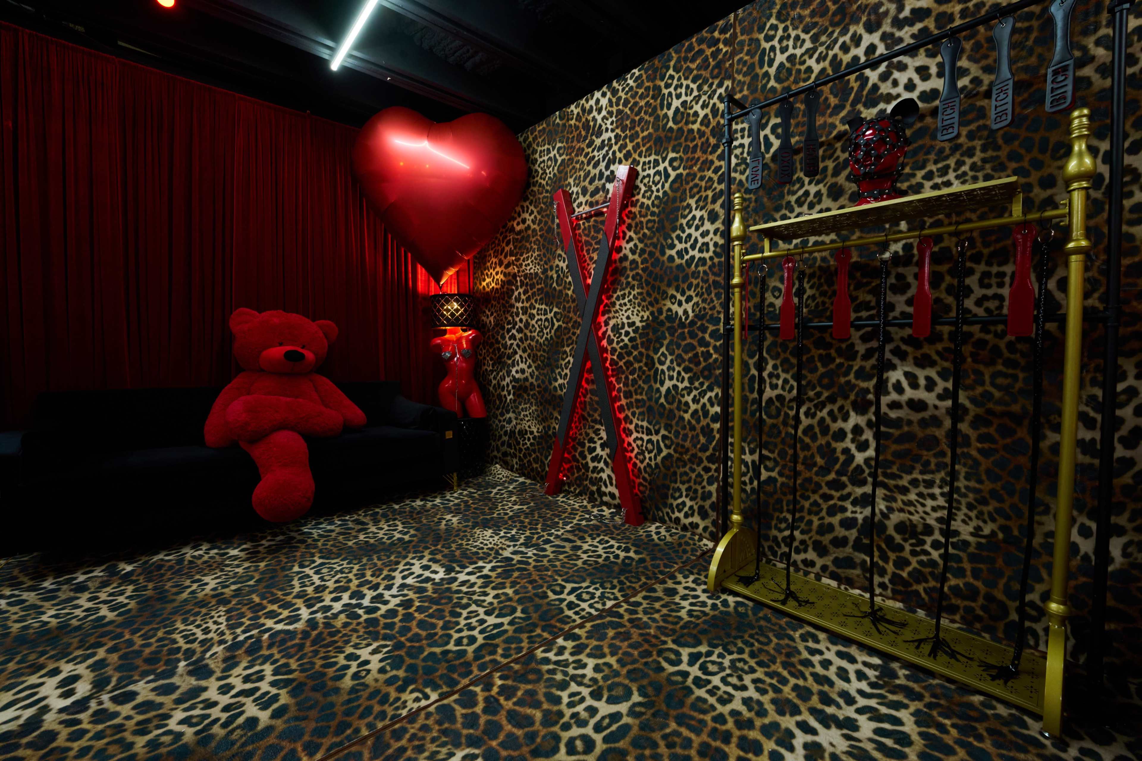 The scene features a room with a leopard-print floor, red walls, a large red heart decoration, and various red accessories including a plush bear and a collection of red items displayed on a gold stand.