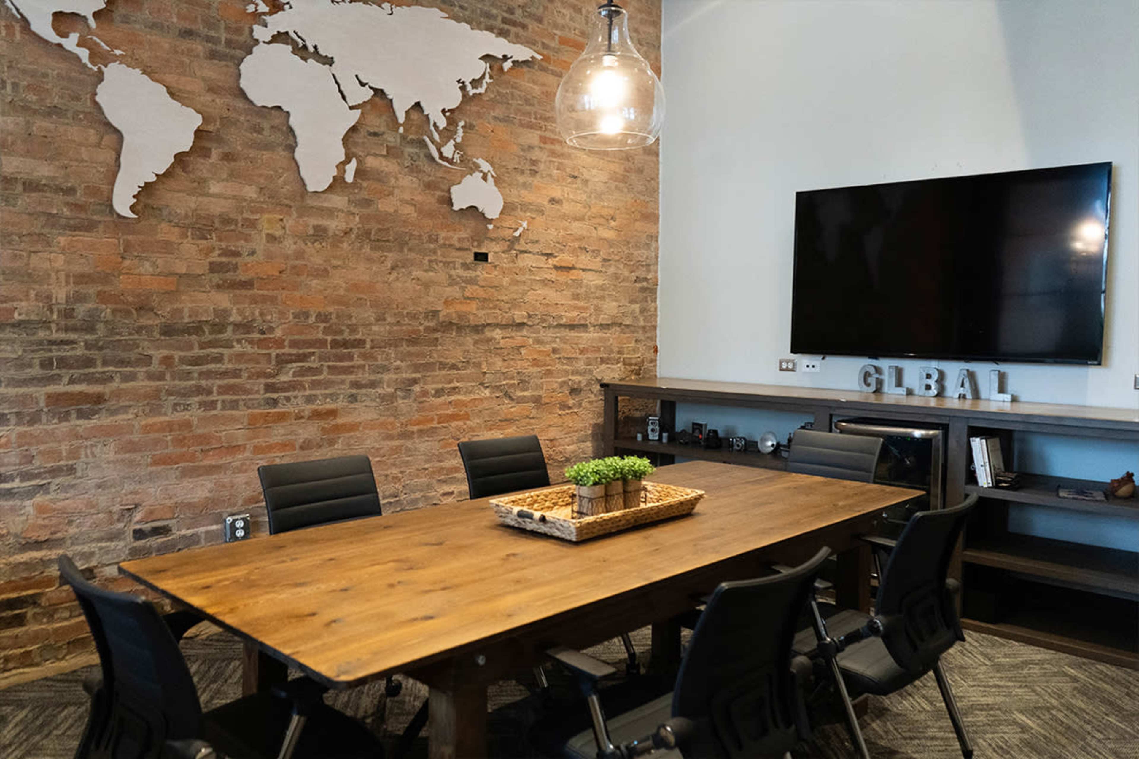 A conference room with a large wooden table surrounded by black chairs, a world map on the brick wall, and a television mounted on the adjacent wall.