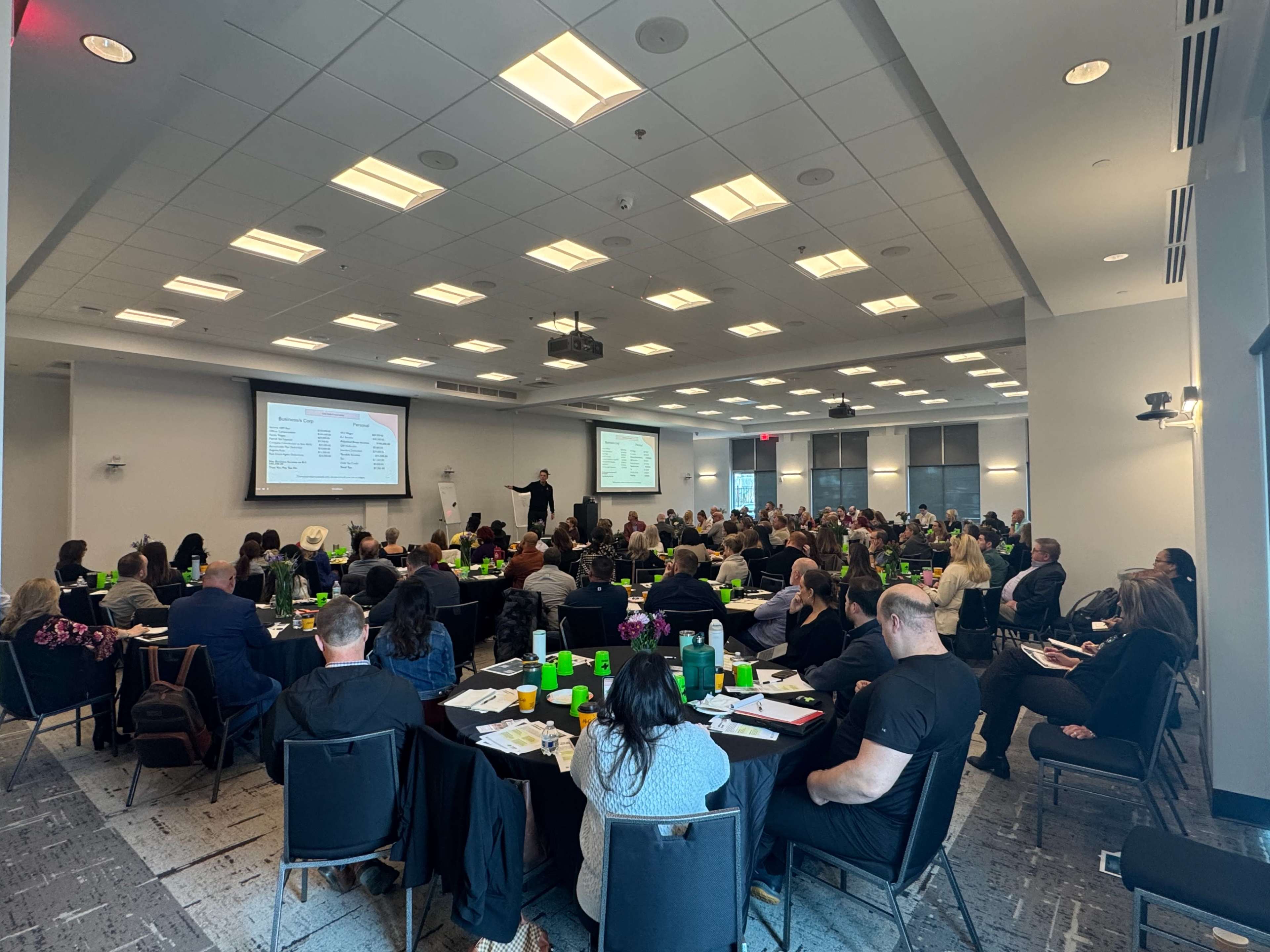 A large group of people sits at tables in a conference room, attentively watching a presenter speaking at the front of the room.