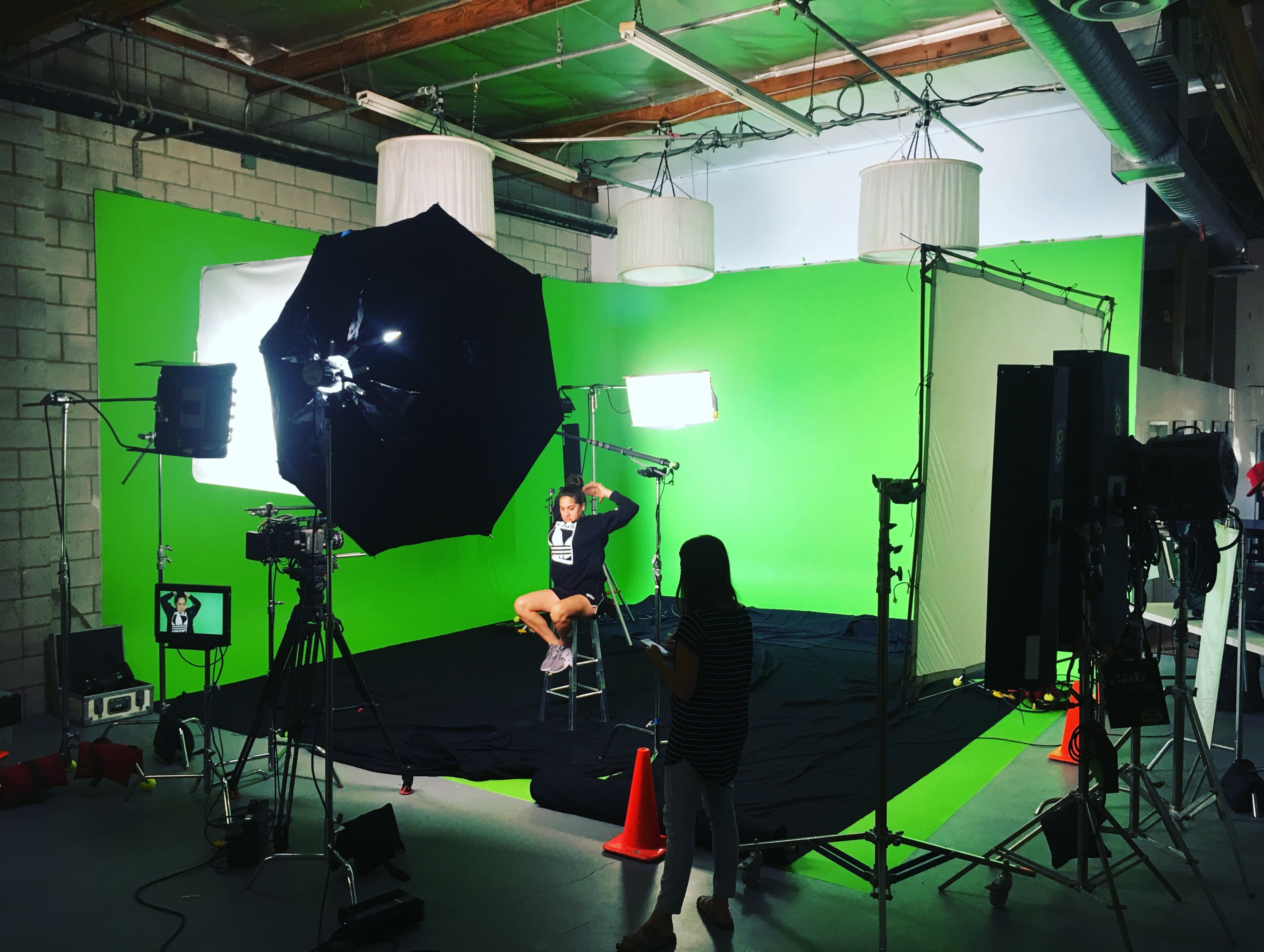 A person sits on a stool in front of a green screen surrounded by various cameras and lighting equipment in a studio setting.
