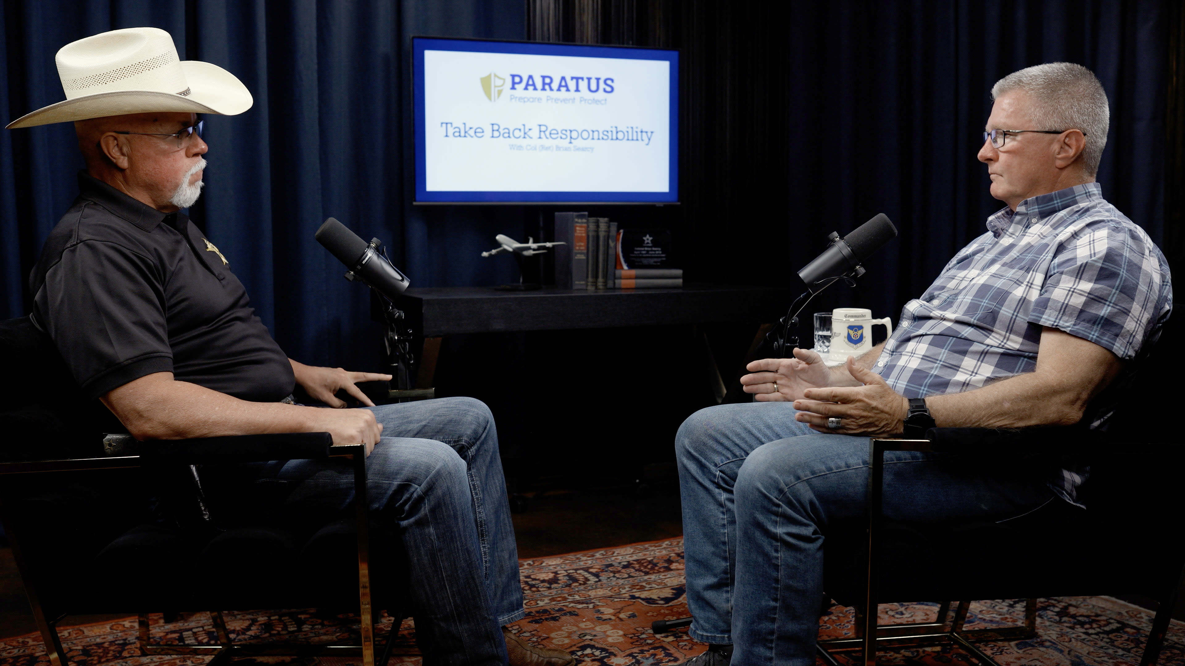 Two men, one wearing a cowboy hat and the other in a plaid shirt, are seated in a studio with microphones, discussing topics related to responsibility, with a sign in the background displaying "Paratus" and the phrase "Take Back Responsibility."