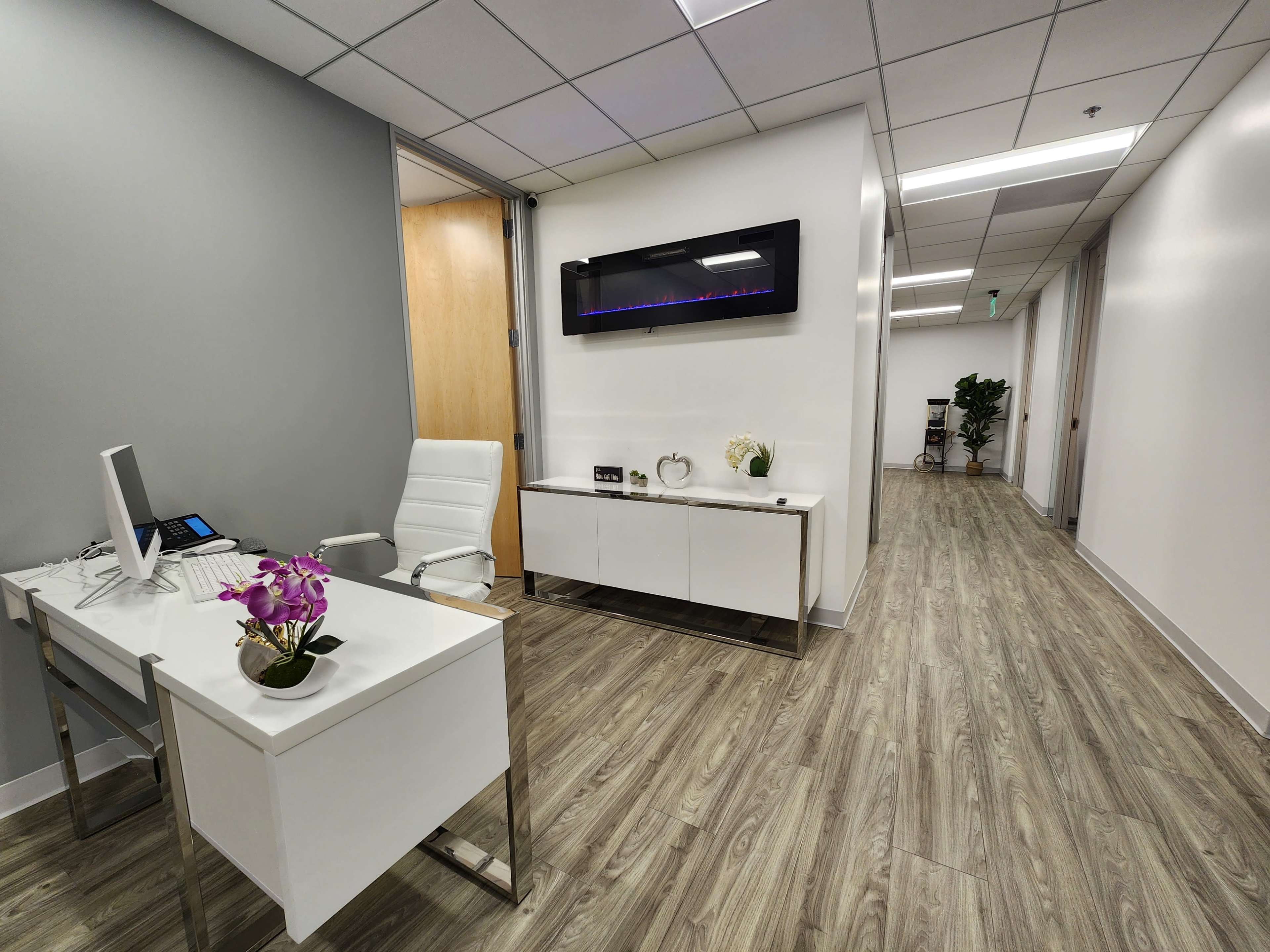 A modern office interior featuring a sleek desk with a computer, a white chair, a wall-mounted television, and a decorative plant along a brightly lit hallway.