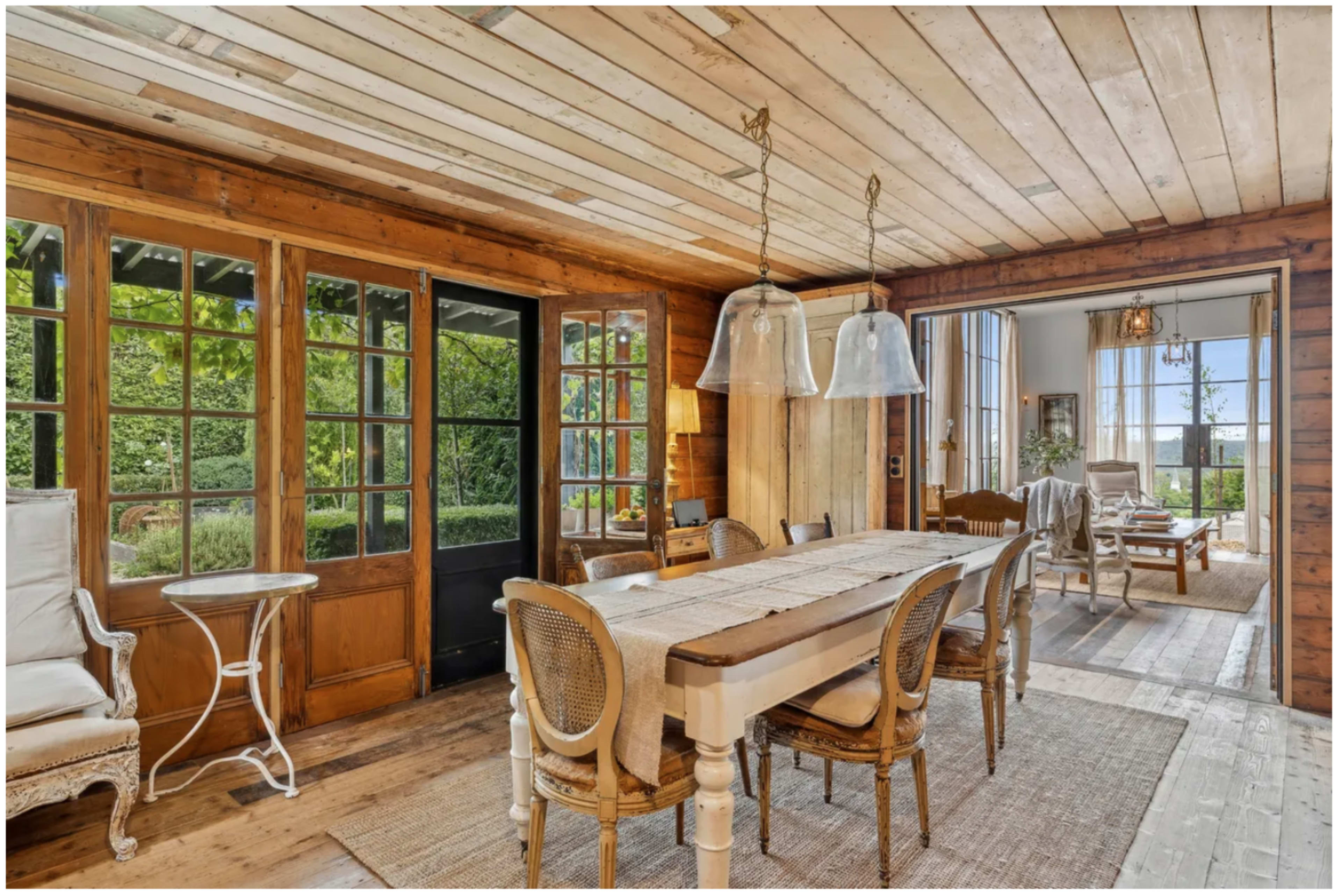 The image shows a wooden-floored dining area with a large table, surrounded by woven chairs, and features large windows that open to a garden view.