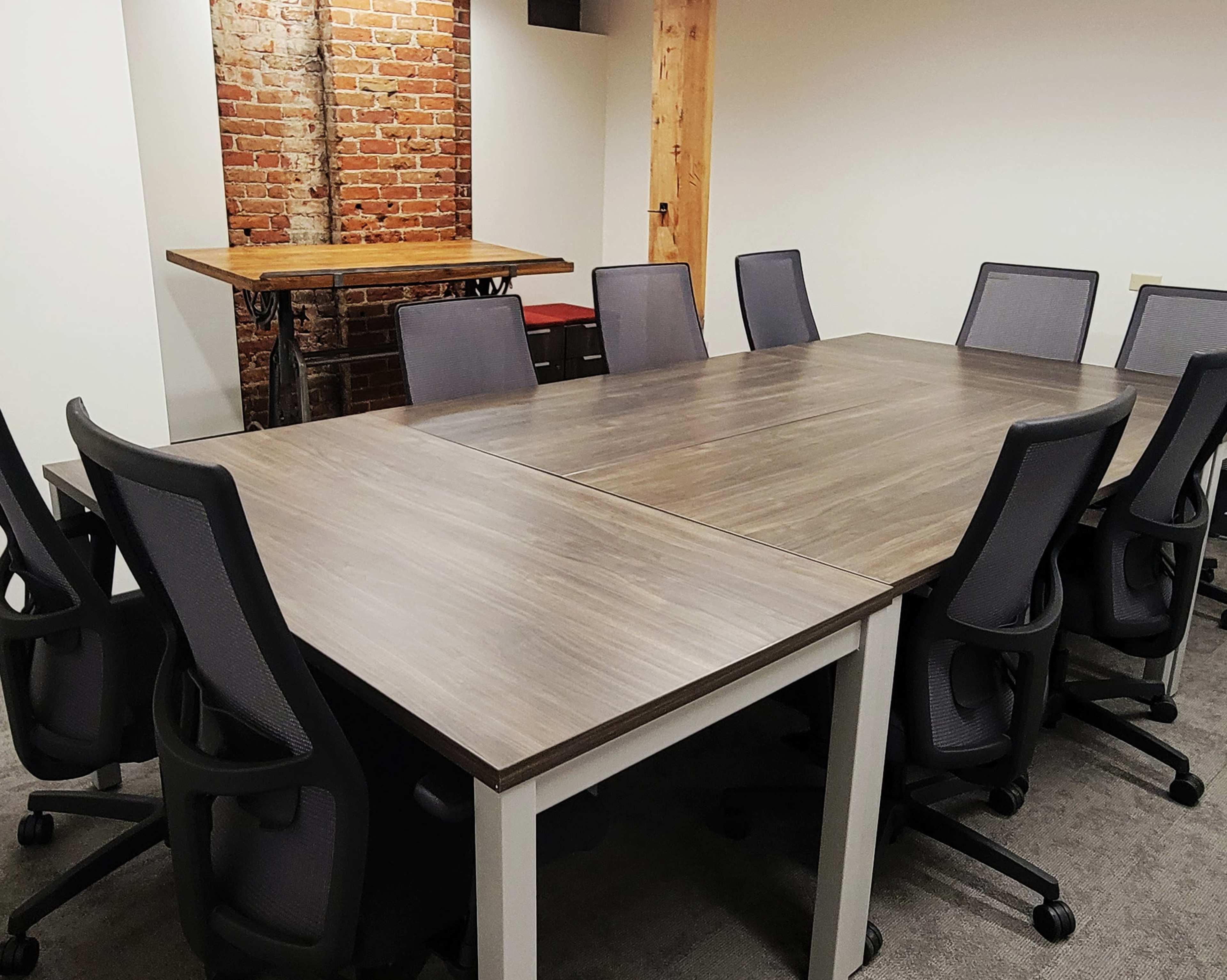 A conference room features a large rectangular table surrounded by office chairs, with a brick wall and a wooden table in the background.