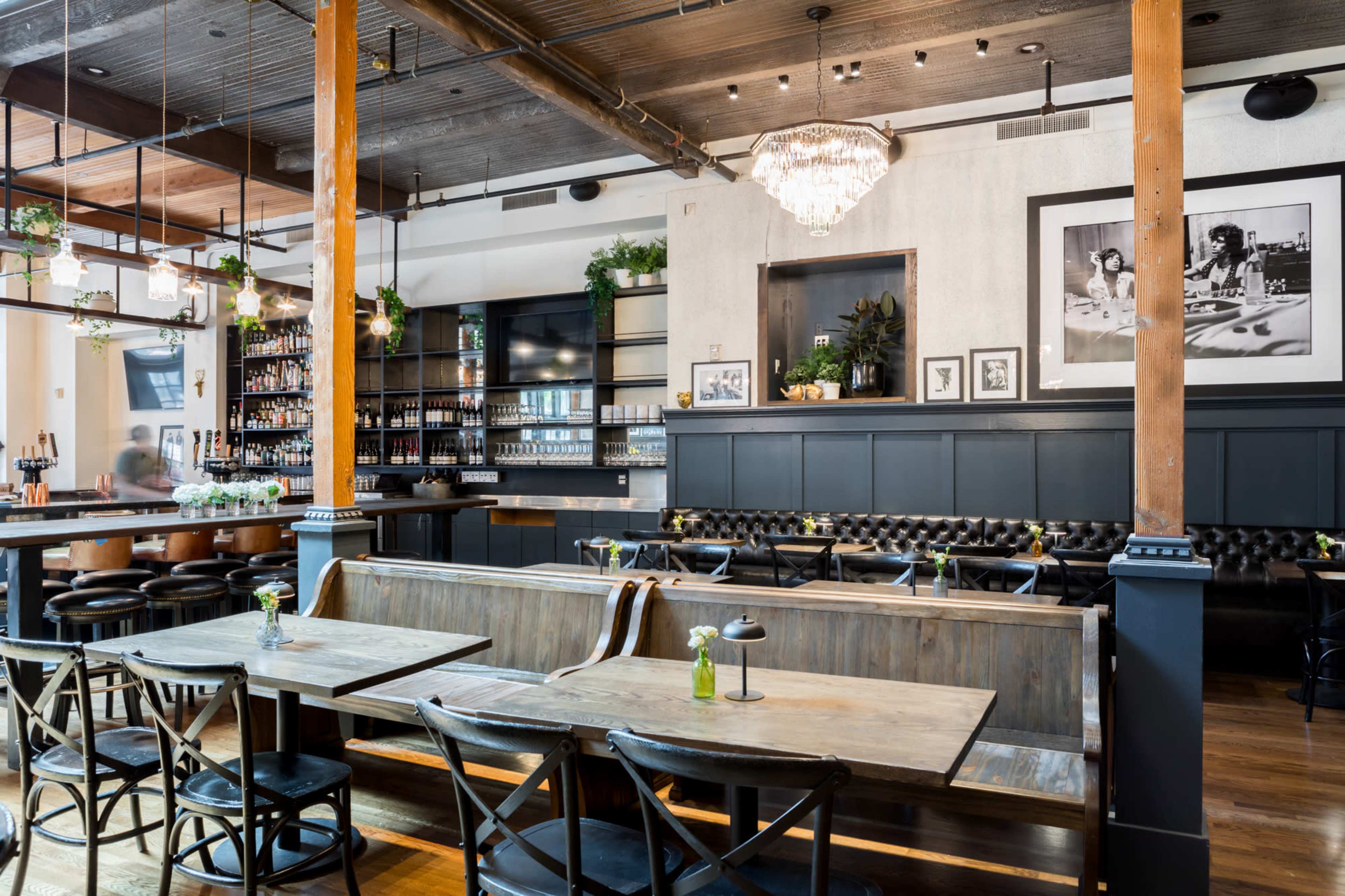 The image shows a stylish restaurant interior featuring wooden tables, black chairs, and a bar lined with drinks, accented by a chandelier and various framed photos on the walls.