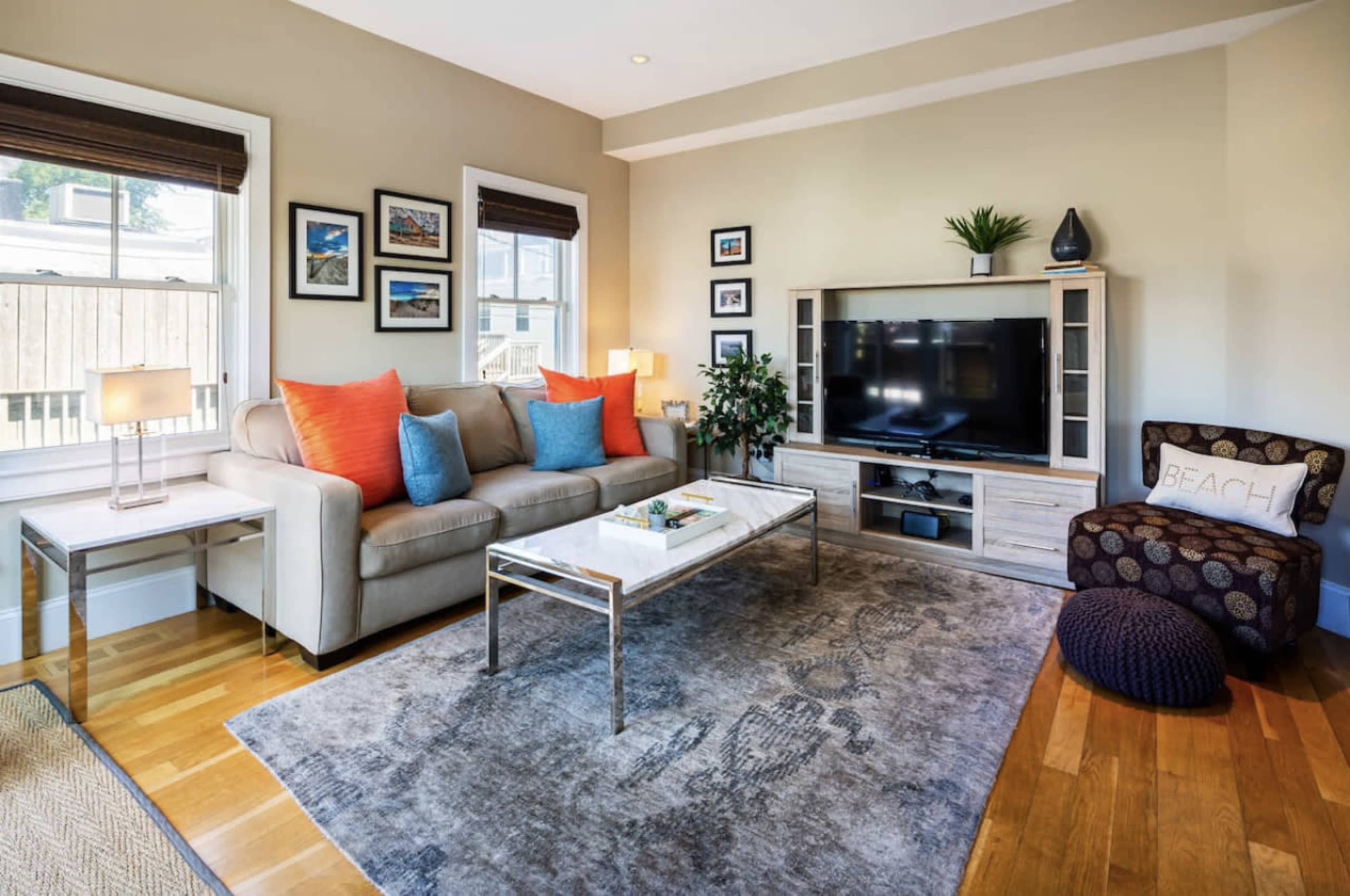 A living room features a light gray sofa with orange cushions, a coffee table, a television unit, and a patterned area rug, all arranged around a wooden floor.