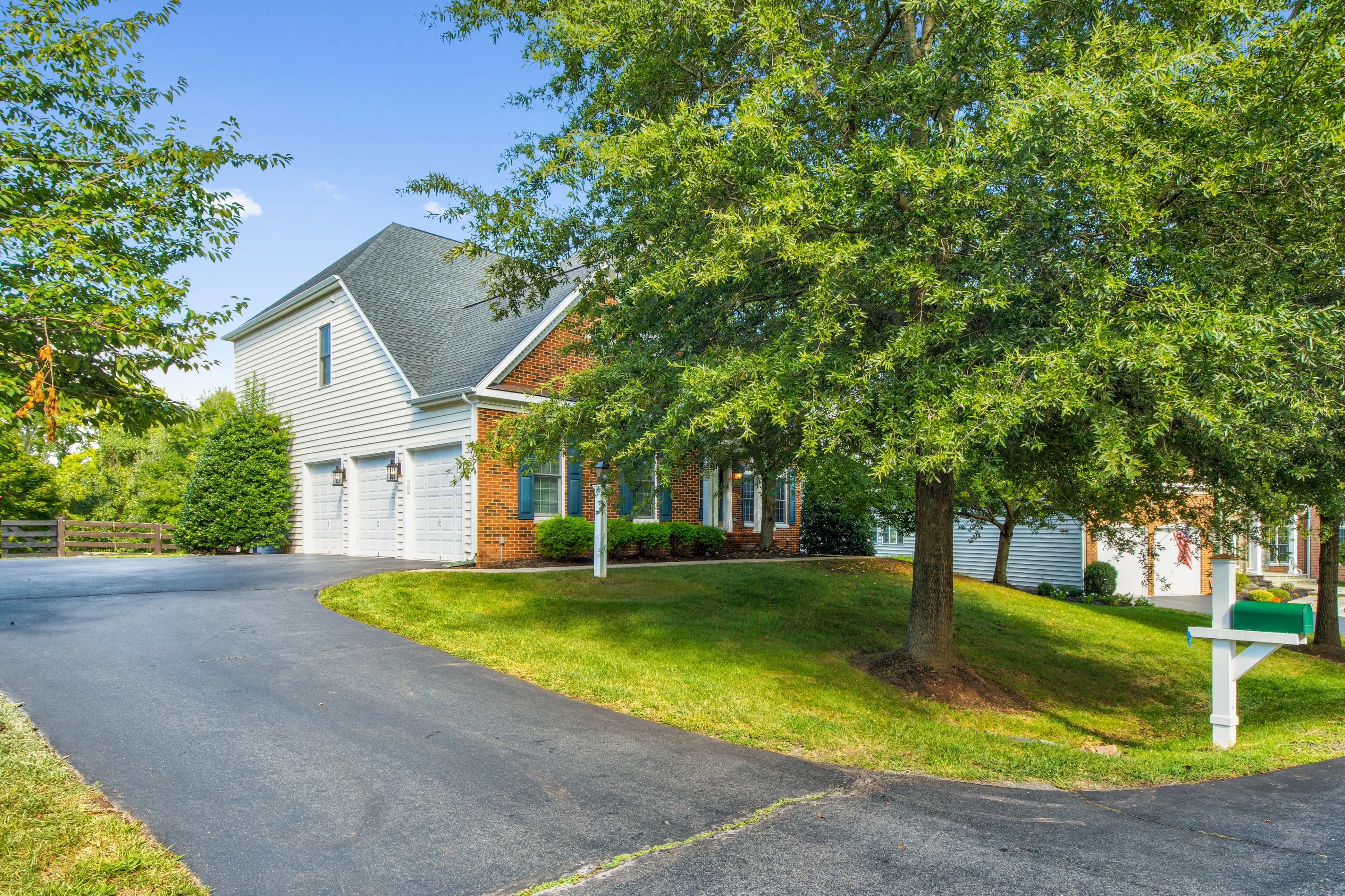 A brick and siding house with a curved driveway and a large tree in the front yard is surrounded by neatly mowed grass.