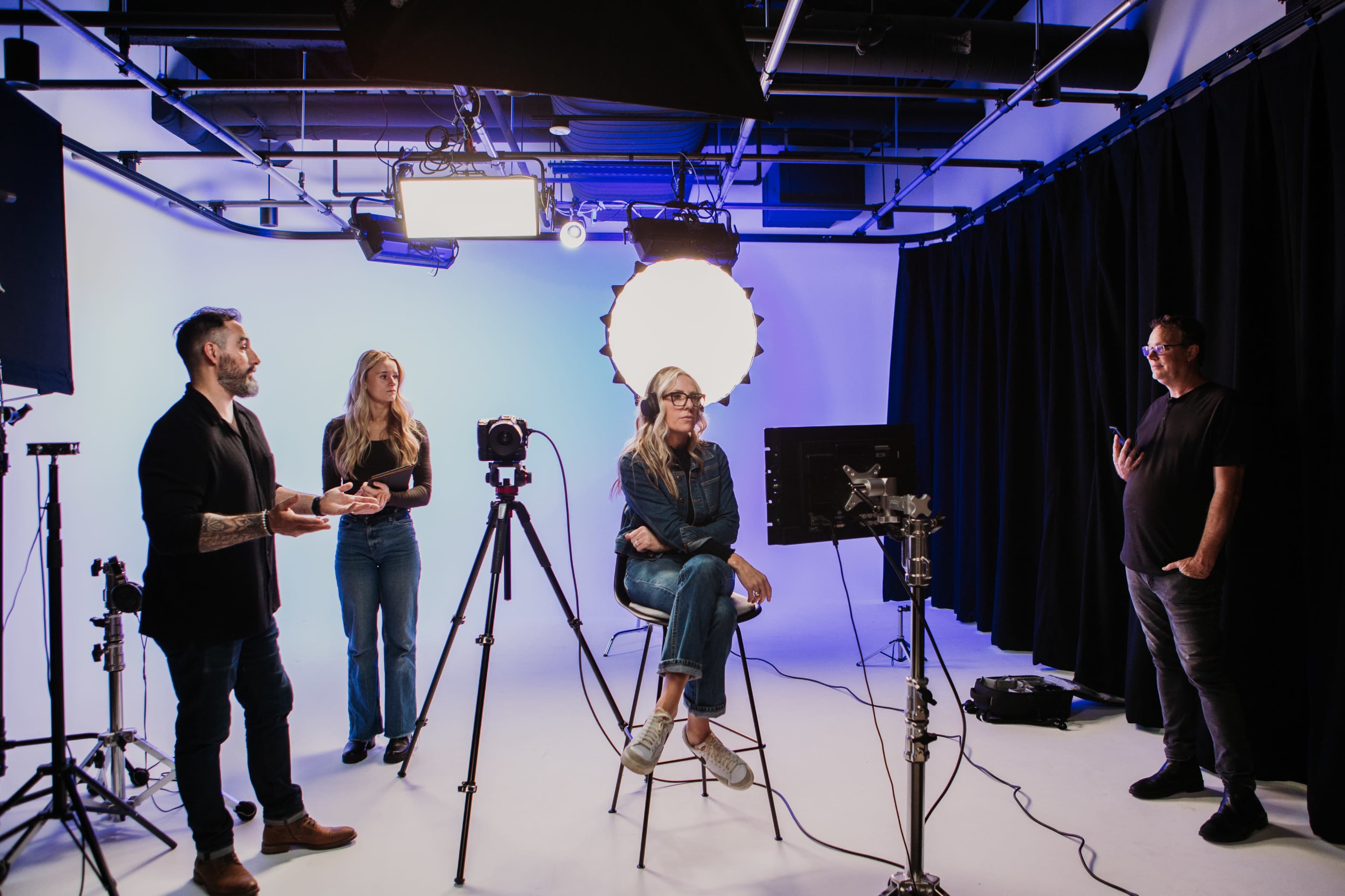 A group of four people is set up in a studio with professional lighting and equipment, while one person sits on a stool in front of a camera.