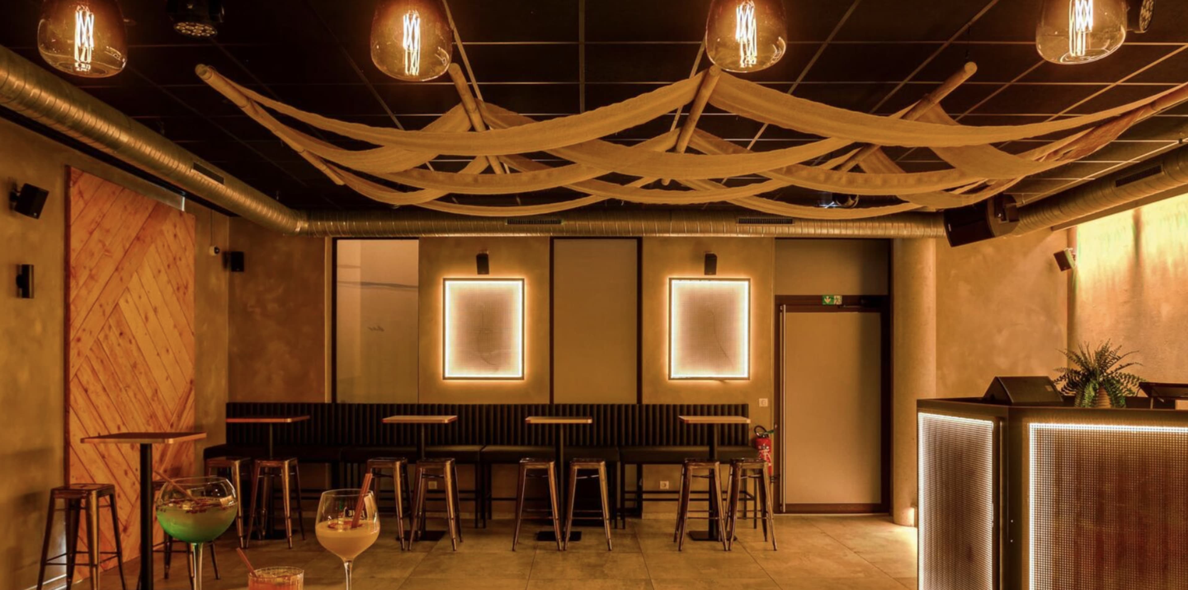 A modern bar interior features minimalistic decor with wooden seating, illuminated frames, and ceiling drapes.