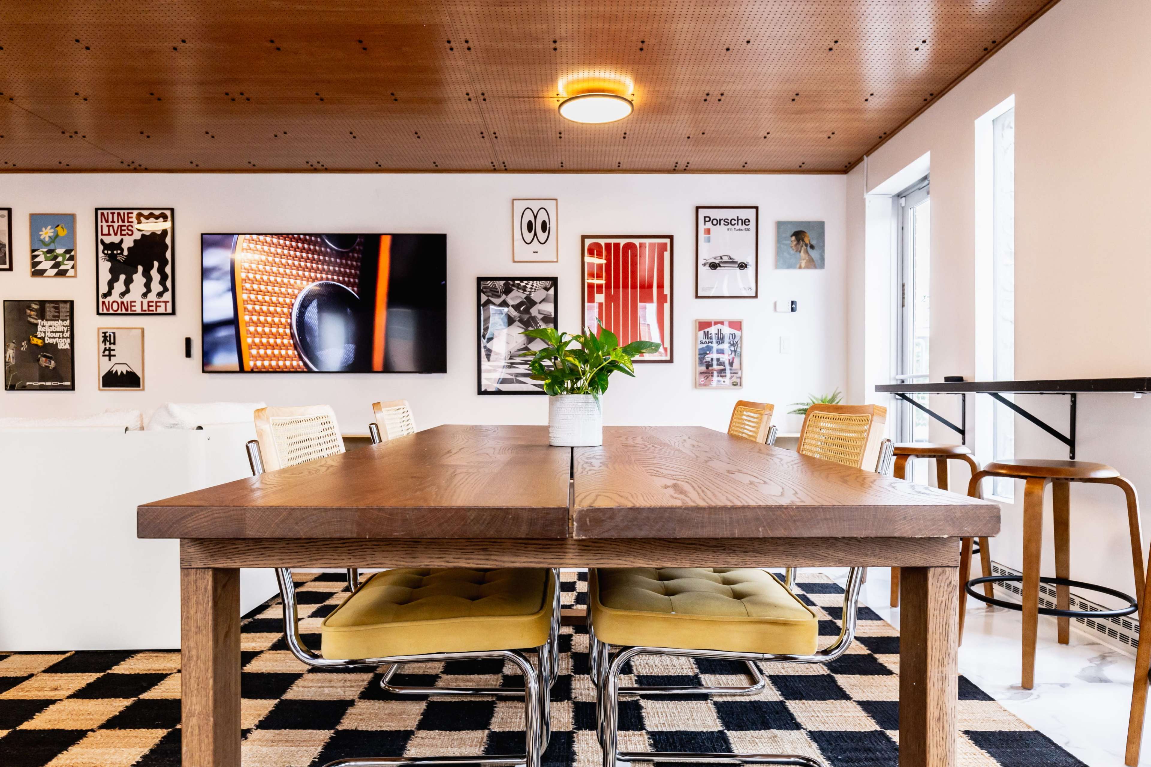 A wooden dining table with yellow chairs is set in a well-lit room featuring various framed artworks on the walls and a large television.