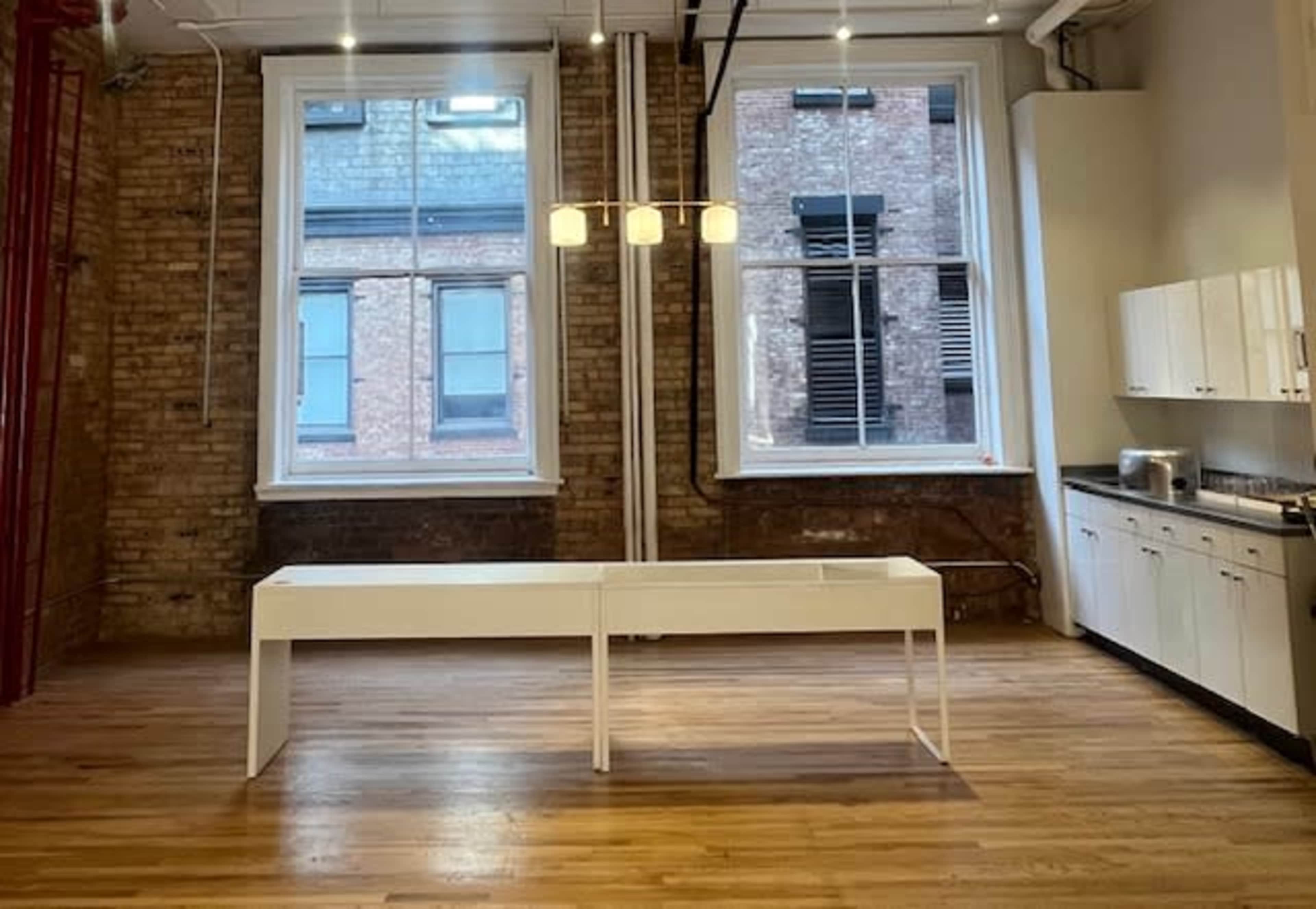 The image shows a spacious room with exposed brick walls, large windows, and a white table in the center, accompanied by a modern light fixture above.