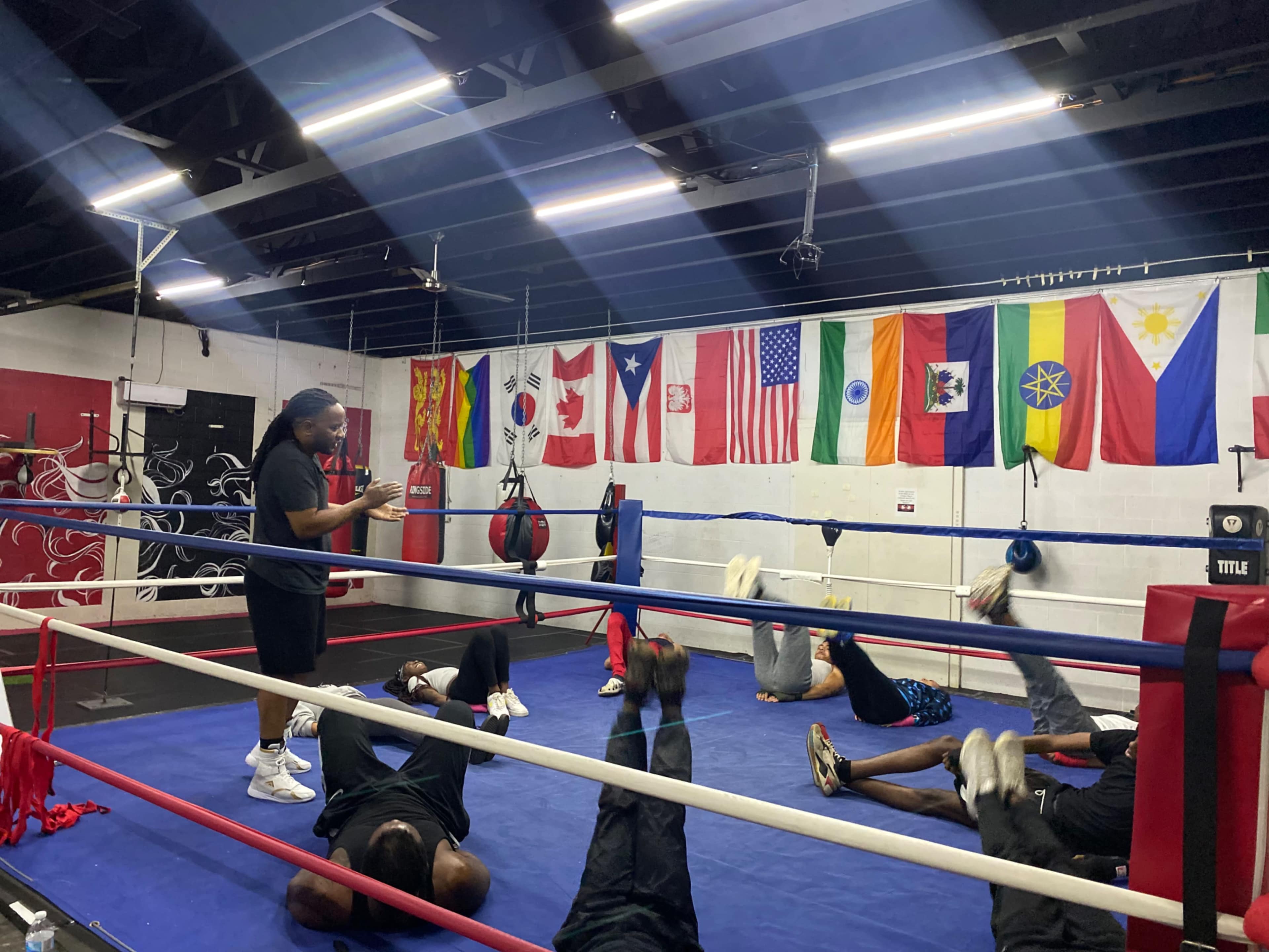 A boxing coach instructs a group of individuals performing exercises on the mat in a gym surrounded by various national flags.