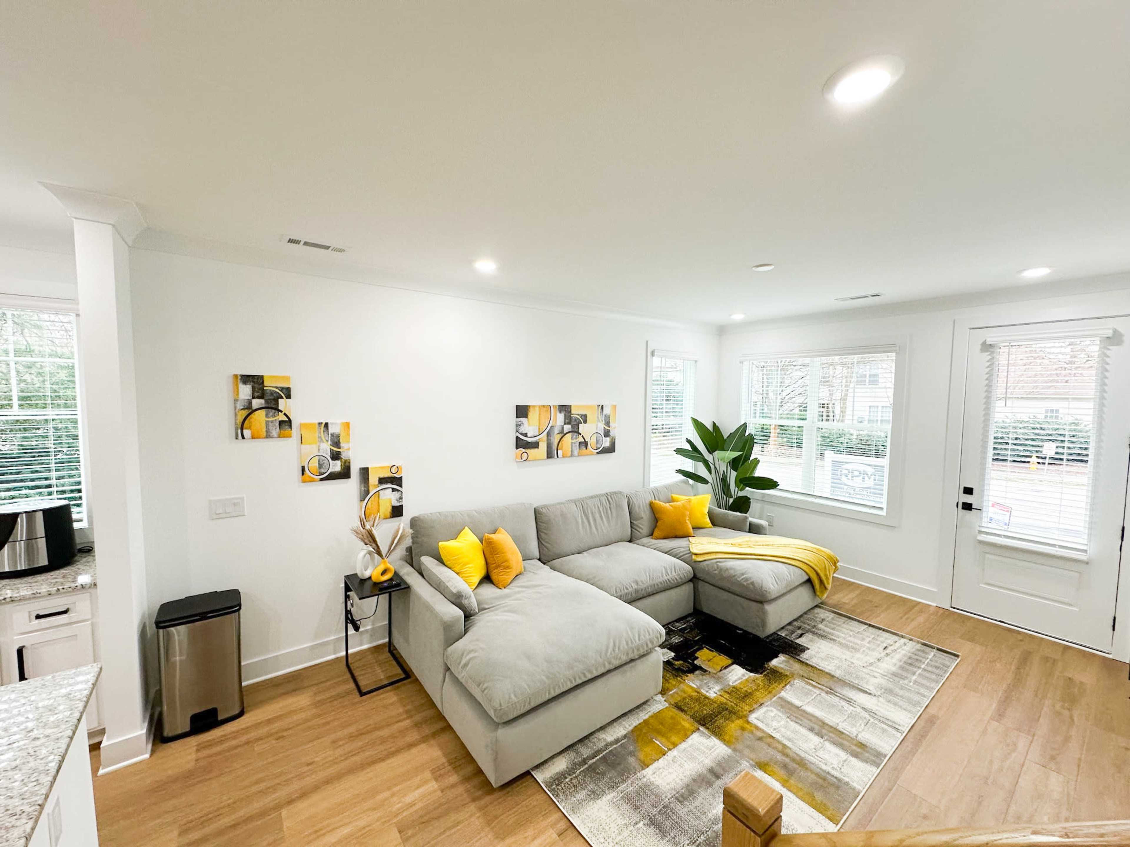 The living room features a large gray sectional sofa adorned with yellow pillows, a rug with geometric patterns, and a potted plant by the window.