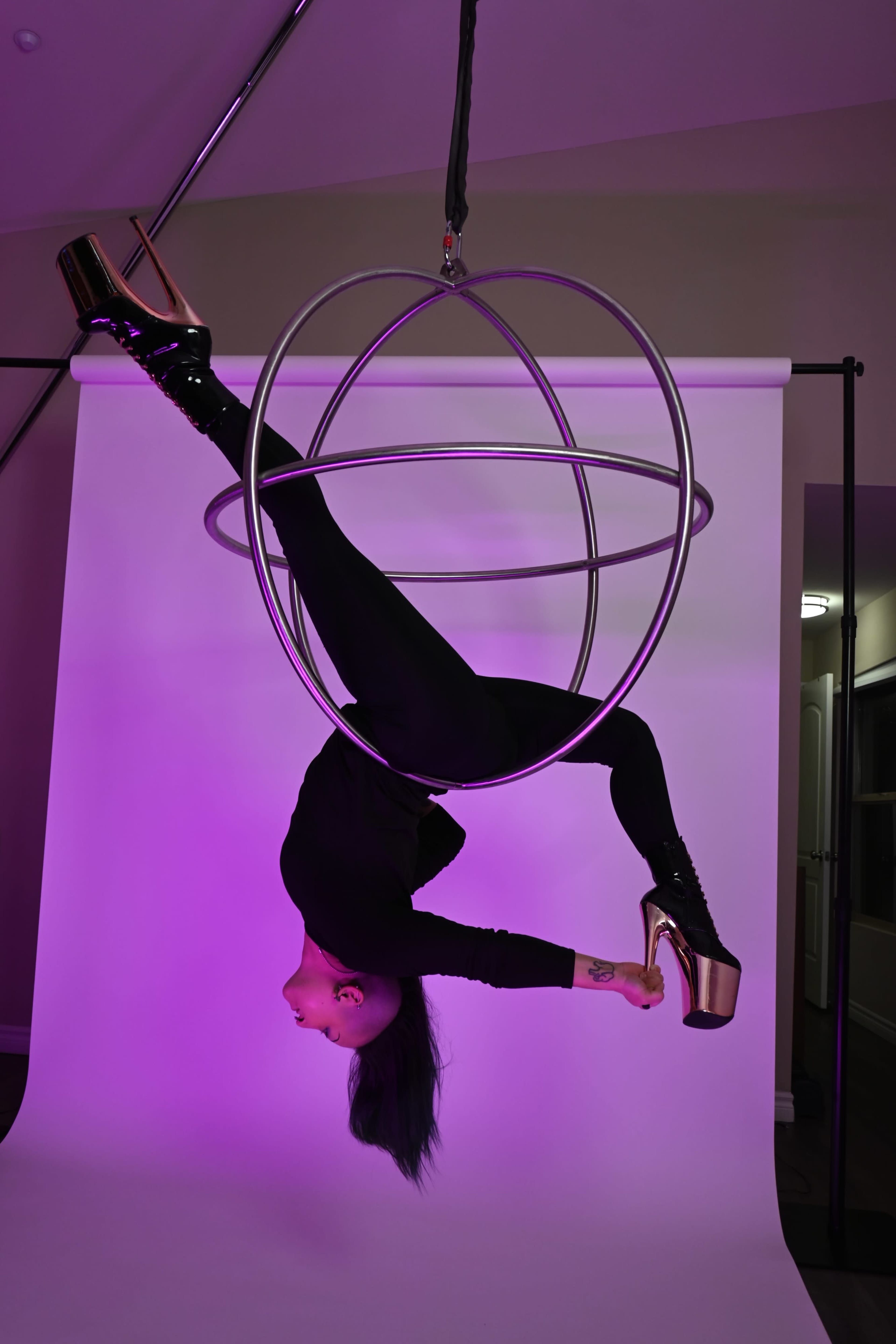 A performer in a black outfit and high heels is suspended upside down in a metallic hoop against a purple-lit backdrop.