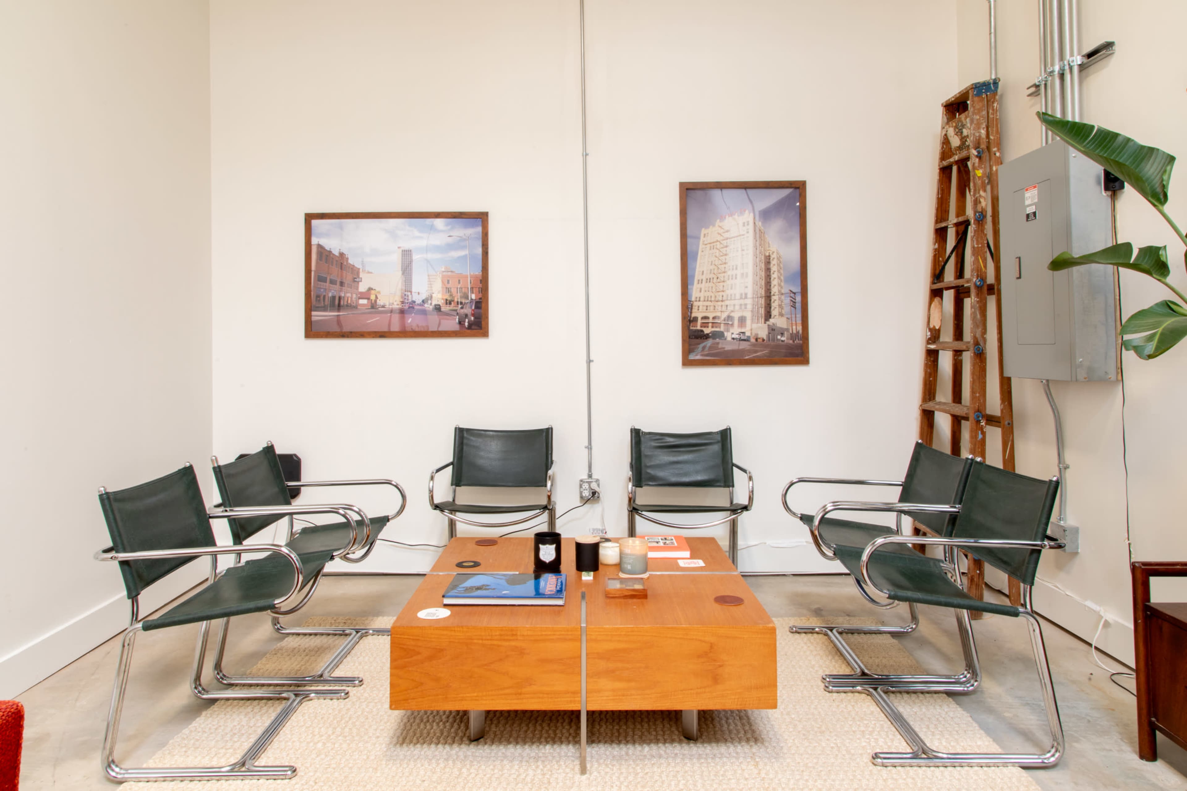 A waiting area with six green chairs arranged around a wooden coffee table, featuring two framed pictures on the walls and a ladder leaning against one side.