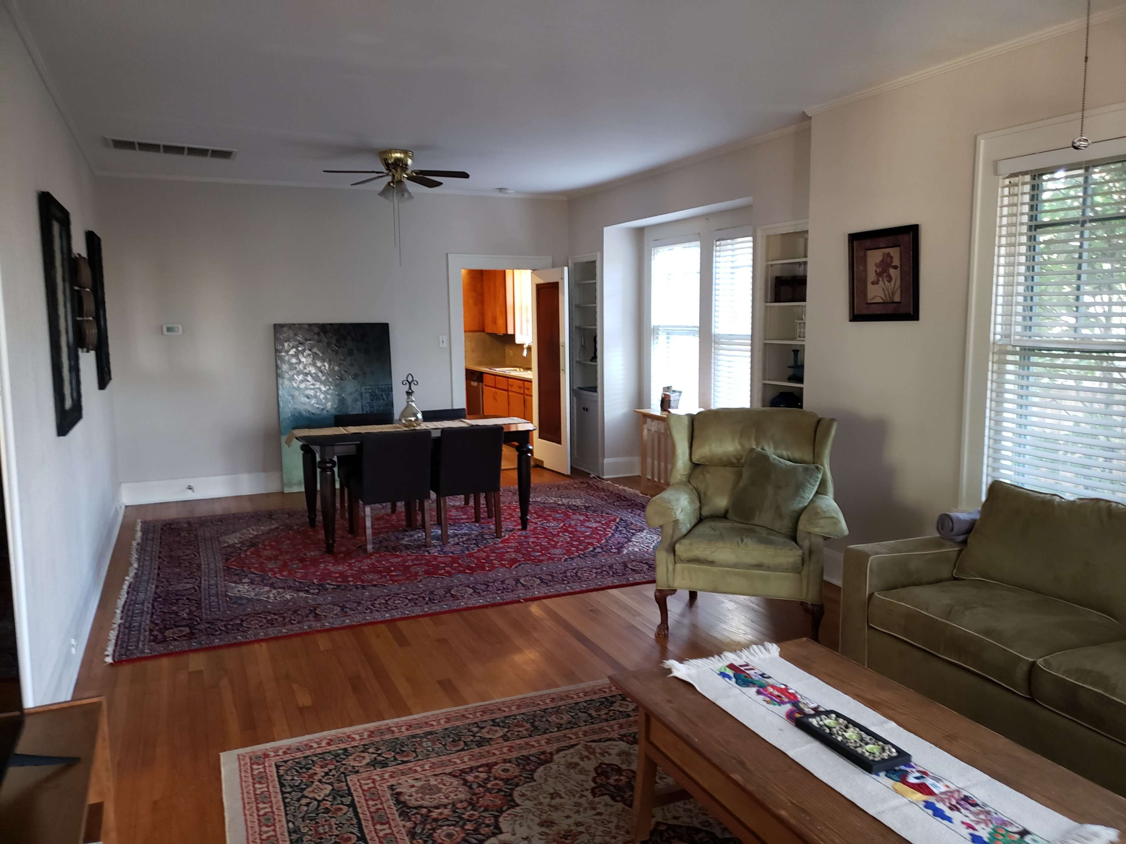 The image shows a living room with a dining table set for four, a green armchair, and a rug on wooden flooring, with a kitchen visible in the background.