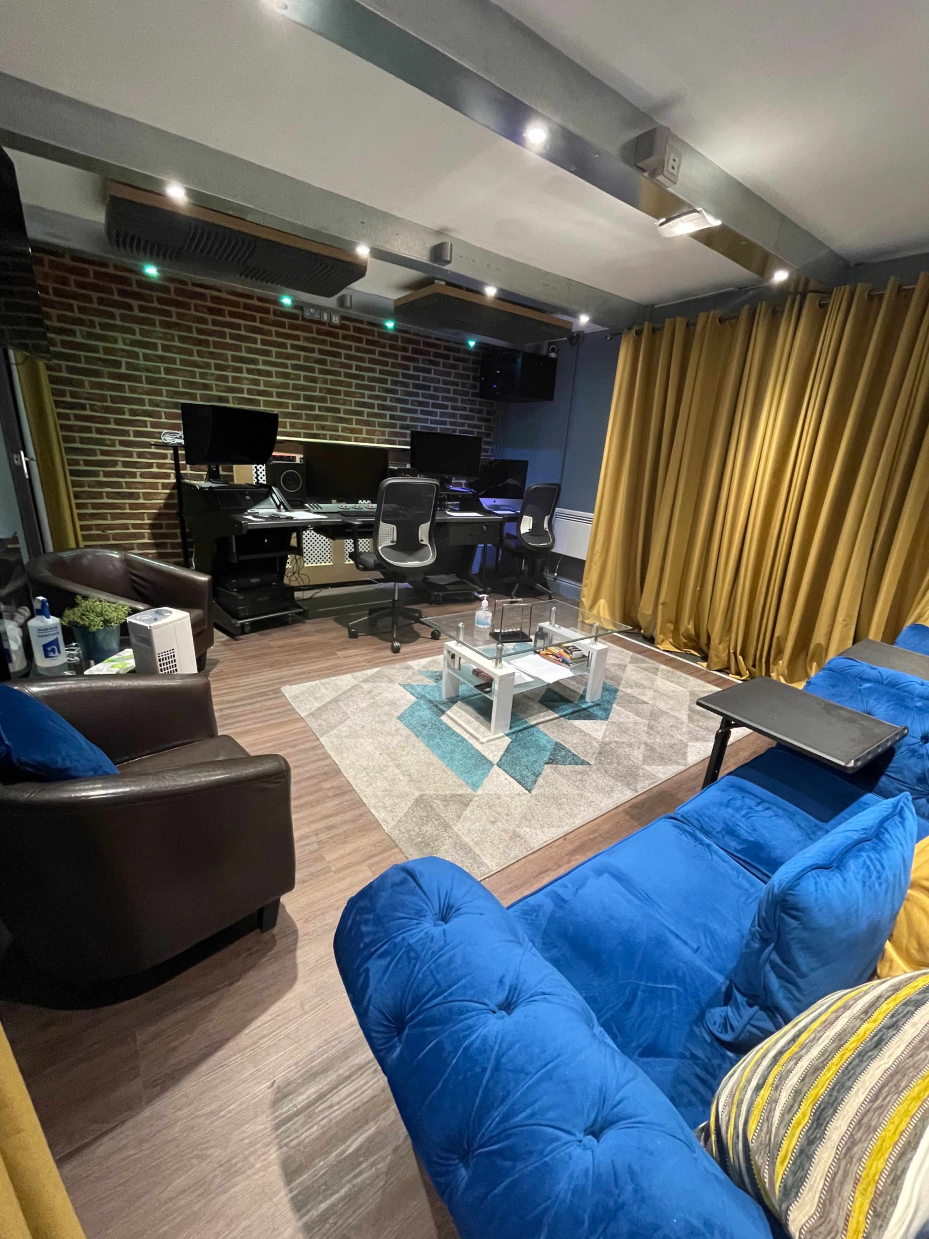 A modern recording studio with a brick wall, dark furniture, a blue couch, and curtains, featuring multiple computer screens and a coffee table in the center.